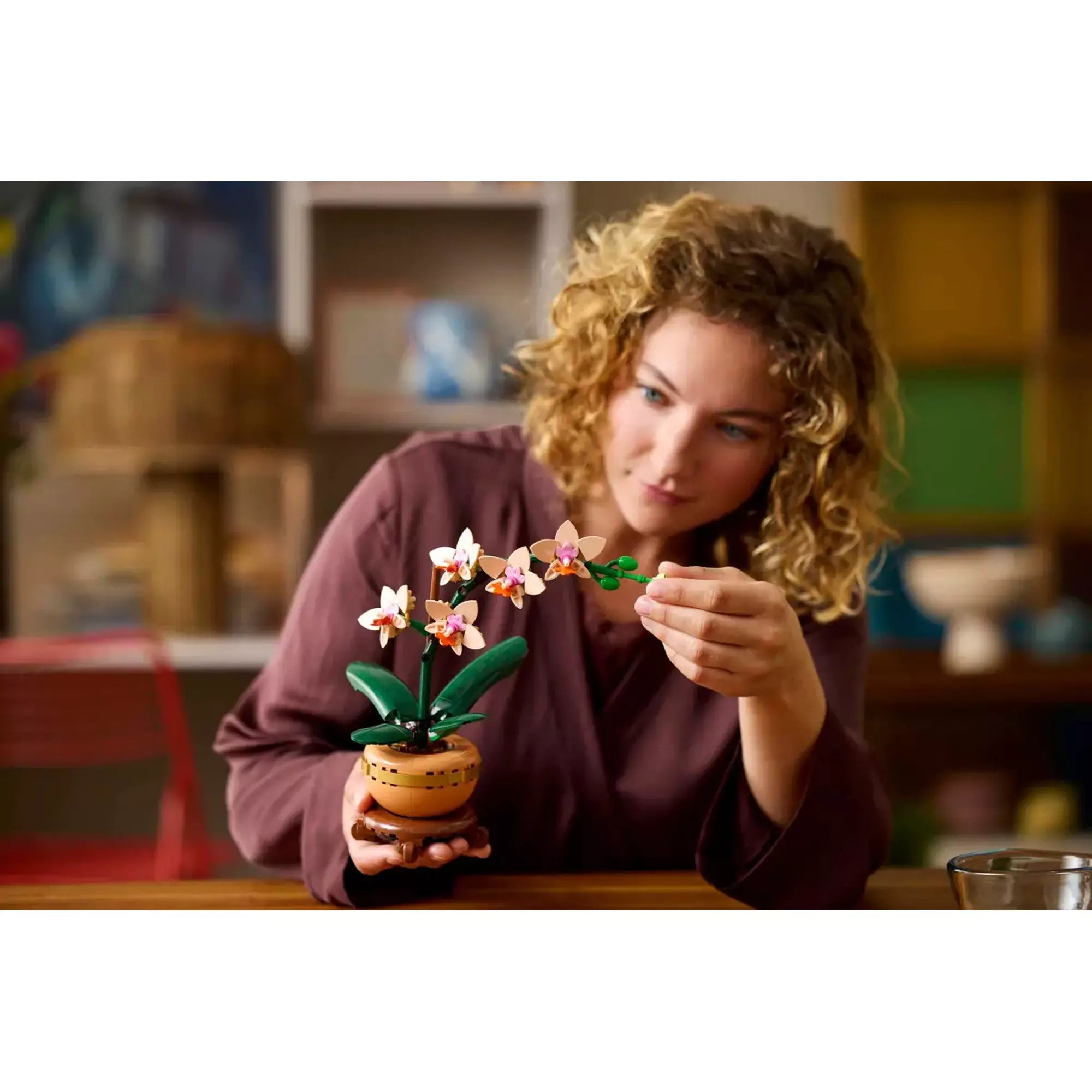 Egy nő kézben tartja és finoman igazítja a LEGO Mini Orchidea (10343) virágait, miközben a részleteket csodálja