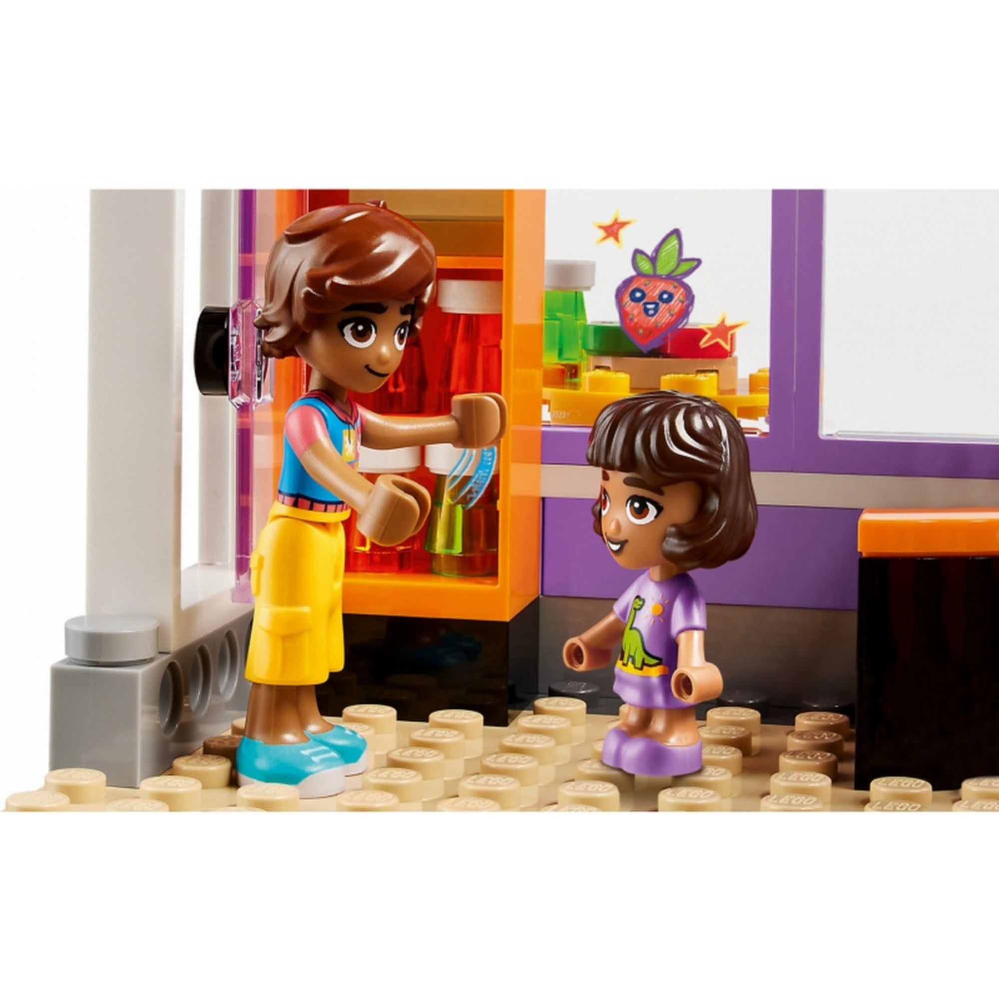 Leo átad egy sütit Albának a LEGO Friends közösségi konyha épületében, színes háttérdekorációval és játékos részletekkel.