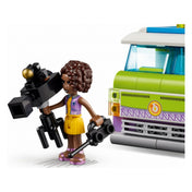A LEGO Friends egyik szereplője, Aliya, sárga ruhában, kezében egy profi videókamerával, a híradós furgon zöld-fehér elejének közelében áll.