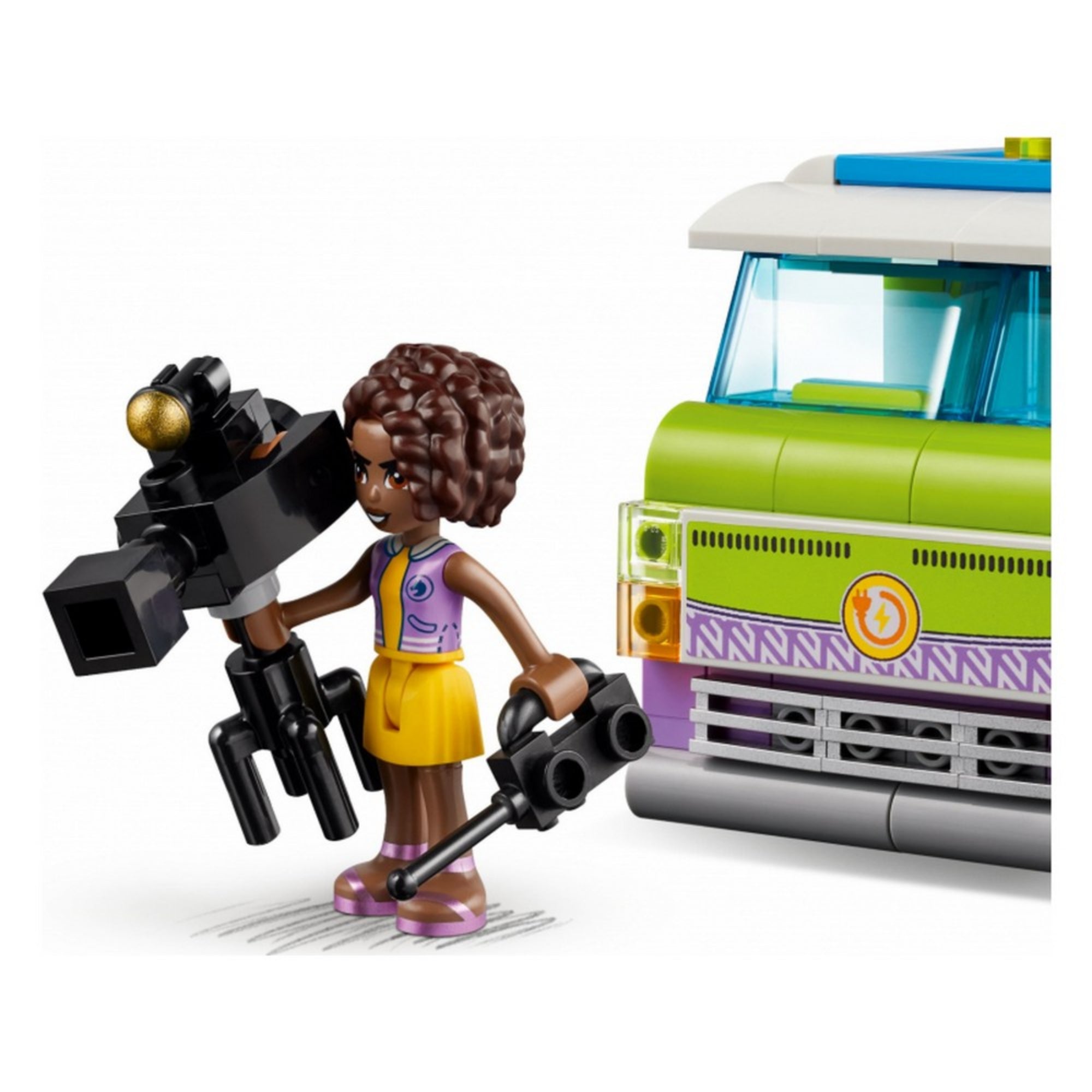 A LEGO Friends egyik szereplője, Aliya, sárga ruhában, kezében egy profi videókamerával, a híradós furgon zöld-fehér elejének közelében áll.