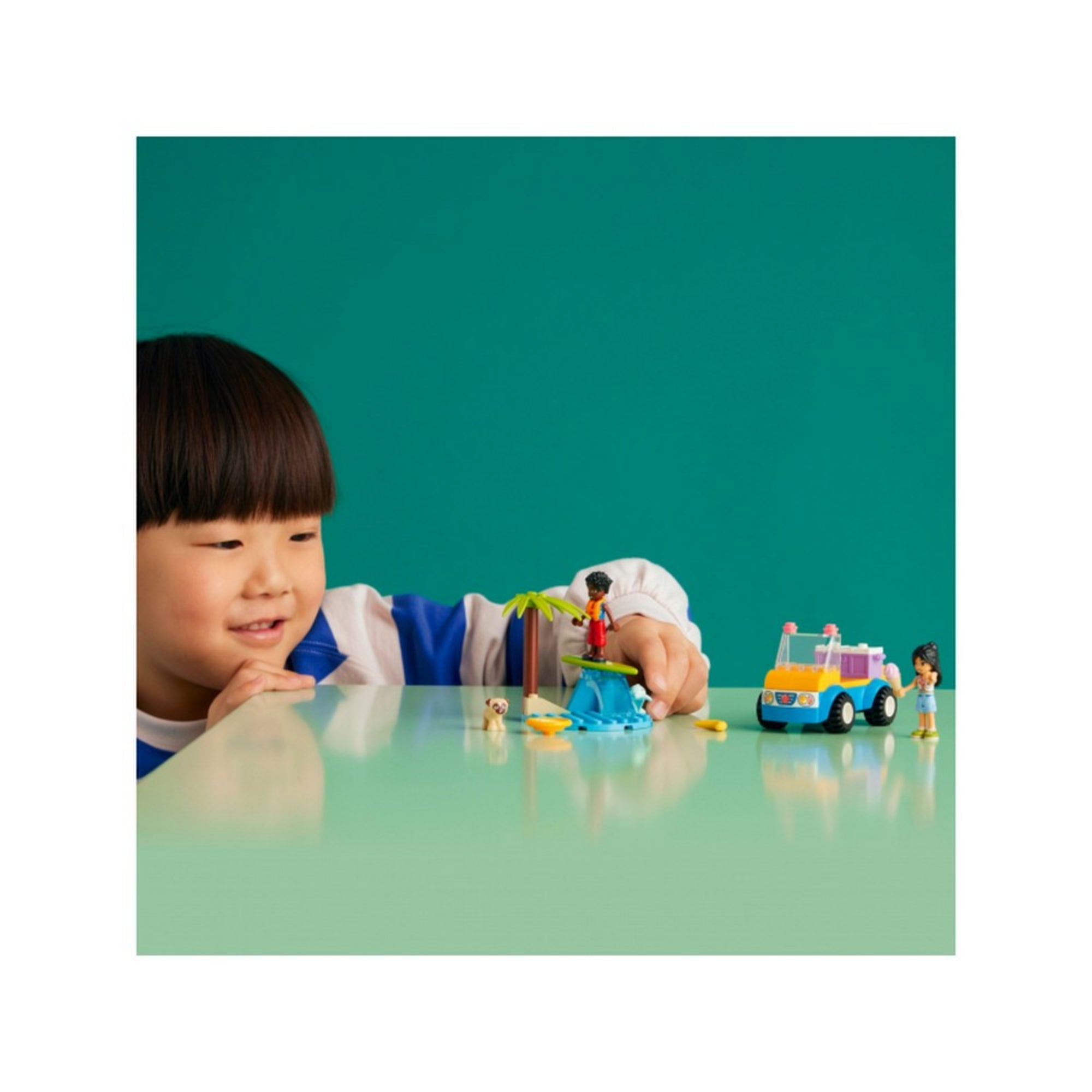 Egy kisfiú örömmel játszik a LEGO Friends Homokfutó móka (41725) készlettel. Az asztalon Liann homokfutóval, Zac szörfözve, a kiskutya pedig a közelben figyel. A háttér letisztult, zöld színű, így a jelenet még jobban kiemelkedik.