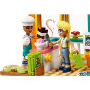 LEGO Friends 41754 Leo szobája – Leo cukrászsapkában egy tejszínhabos édességet, Olly pedig fagyis tálat és kanalakat tart a kezében. A háttérben színes sütősarok, muffin motívumos hűtő és pult látható, a játékélmény központi témája a közös sütés.