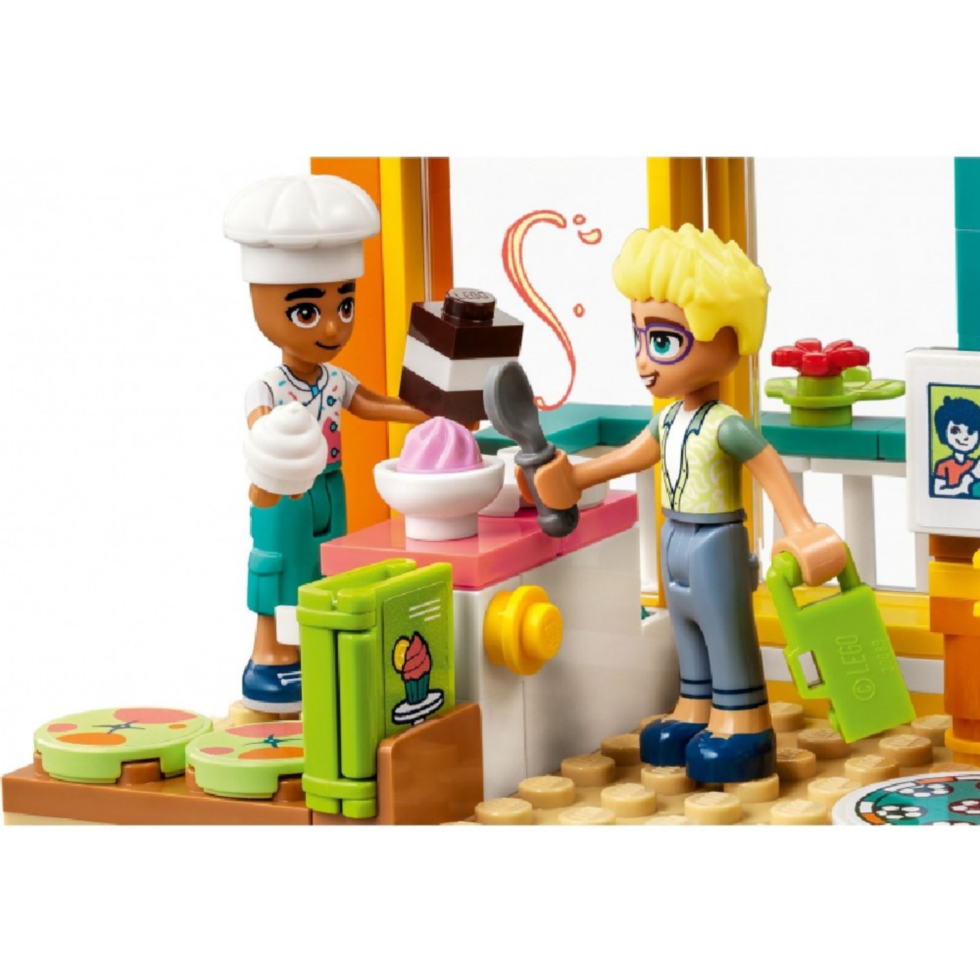 LEGO Friends 41754 Leo szobája – Leo cukrászsapkában egy tejszínhabos édességet, Olly pedig fagyis tálat és kanalakat tart a kezében. A háttérben színes sütősarok, muffin motívumos hűtő és pult látható, a játékélmény központi témája a közös sütés.