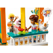 LEGO Friends 41754 Leo szobája – Olly egy pohárral a kezében beszélget Leóval az erkélyen, miközben a fehér macska is csatlakozik hozzájuk. A jelenet hangulatos, virágos erkélyt és beltérből kitekintő nyílászárót ábrázol.