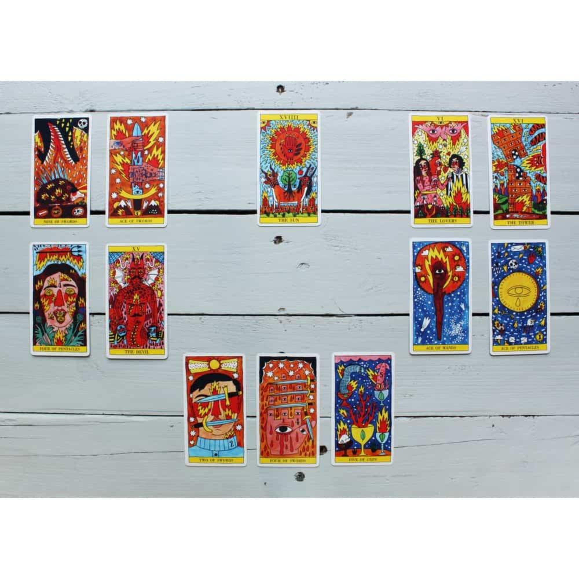 A képen a „Tarot del Fuego” kártyacsomag 14 darabja látható szabálytalan elrendezésben, egy világos, fehérre festett fa felületen. A lapokat Ricardo Cavolo egyedi, szürreális és színpompás stílusa jellemzi: minden kártyán dominálnak a vörös, sárga, kék és narancs árnyalatok. A karakterek gyakran több szemmel, lángoló elemekkel vagy absztrakt szimbólumokkal vannak ábrázolva. Kiemelkedik például The Sun lap, amely középen helyezkedik el, valamint a The Lovers, The Devil és The Tower is.