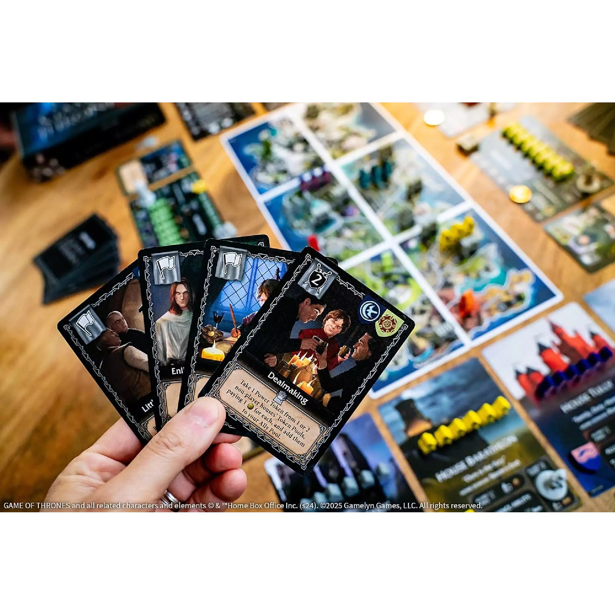 Játékos kézben tartott Tiny Epic Game of Thrones kártyák közelről, köztük Tyrion Lannister karakterkártyája, háttérben a játékmező.