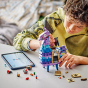 Egy fiú éppen építi a LEGO Fortnite Supply Llama modellt, mellette a tableten a digitális útmutató látható. A képen a játék kreatív építési folyamata és a kiegészítők is megjelennek.