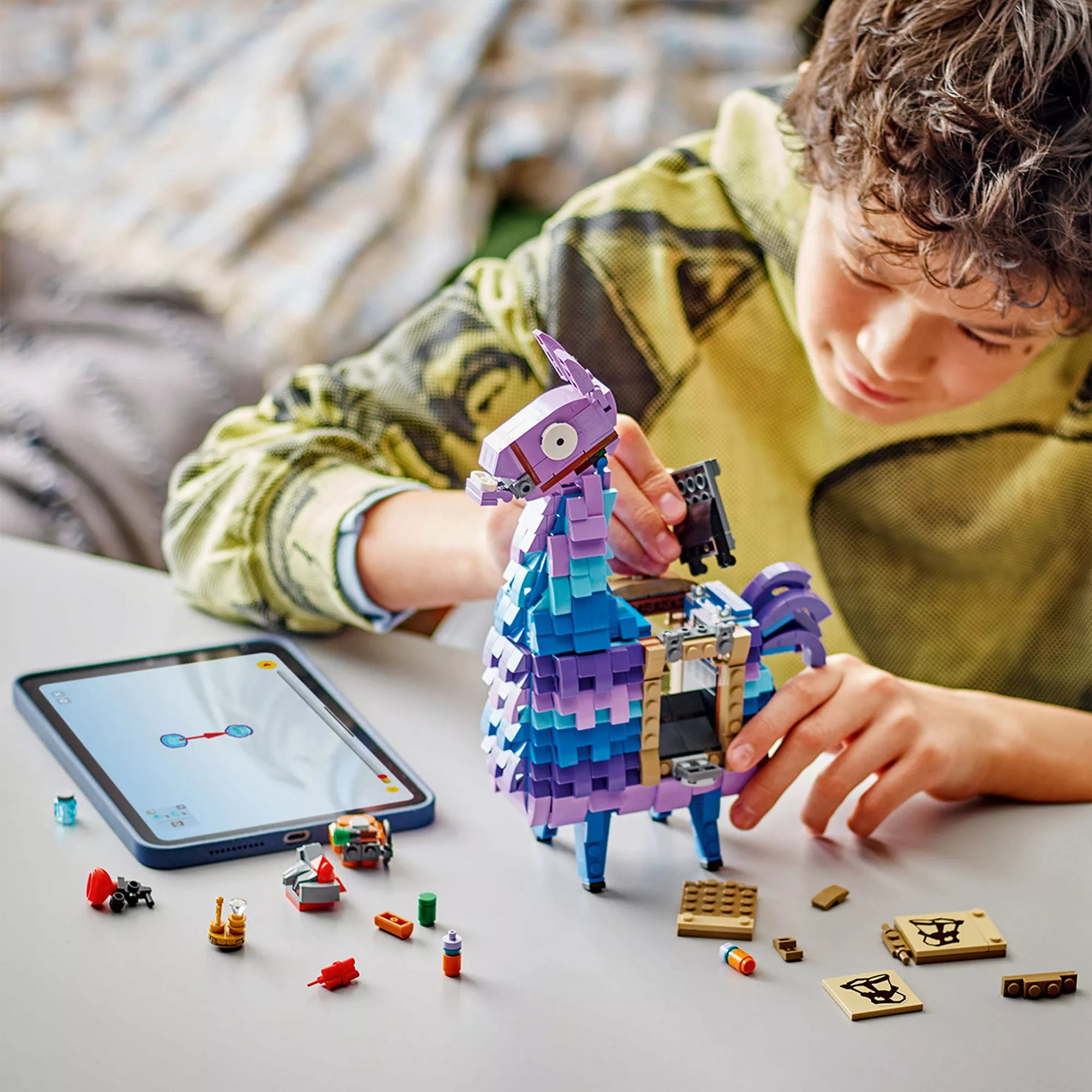 Egy fiú éppen építi a LEGO Fortnite Supply Llama modellt, mellette a tableten a digitális útmutató látható. A képen a játék kreatív építési folyamata és a kiegészítők is megjelennek.