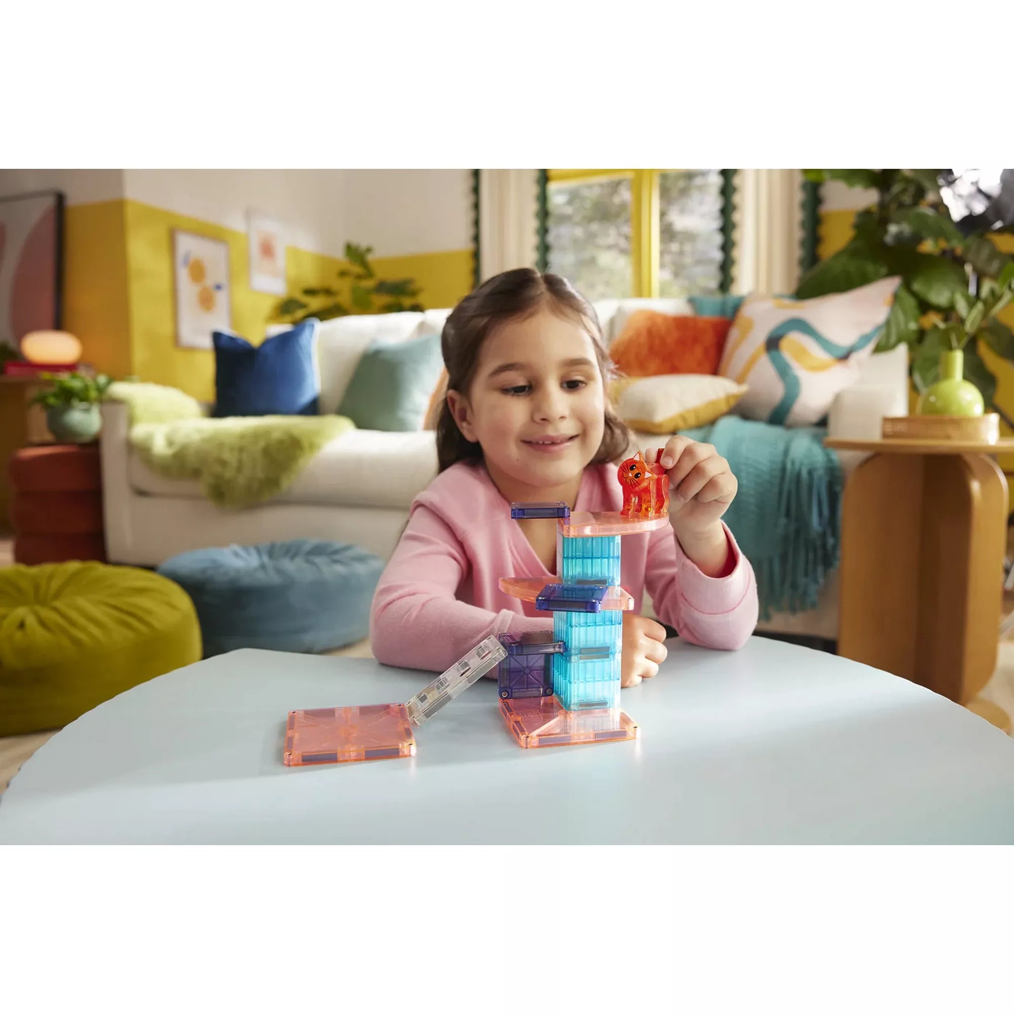 Egy mosolygó kislány játszik a MAGNA-TILES Cat Tree készlettel az asztalon. A gyerek a piros macskafigurát helyezi a színes, mágneses elemekből épített cicatorony tetejére. A háttérben otthonos nappali látható.