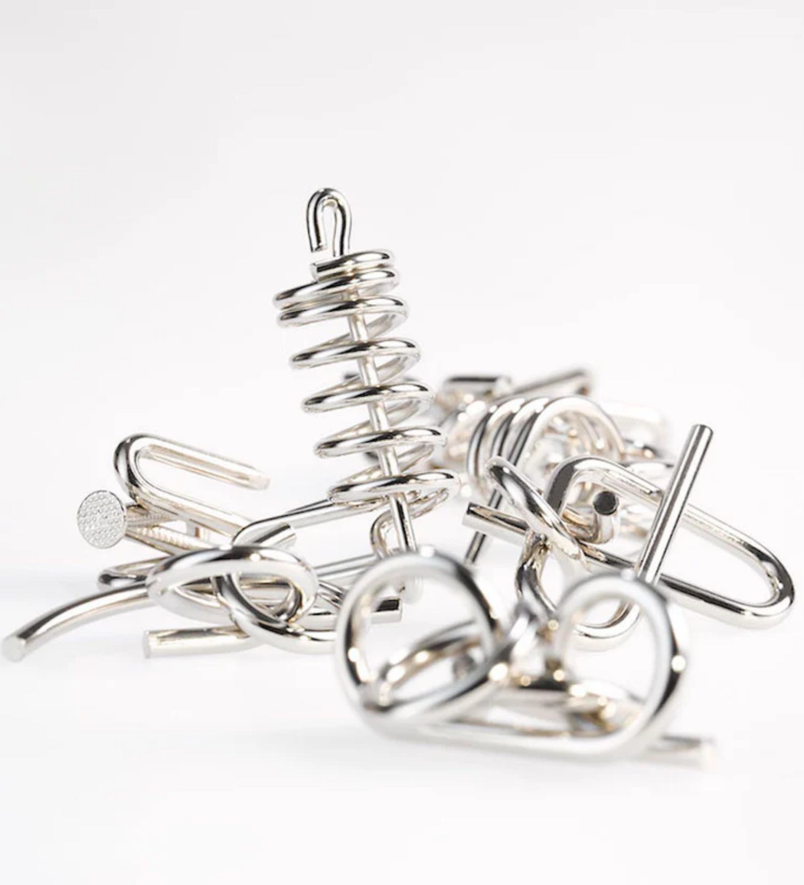 A 10 Metal Puzzle Set zöld változatának tartalma szétszedve, különböző alakú és formájú ezüstszínű fém kirakóelemek halmaza világos háttér előtt, előtérben egy spirálos forma emelkedik ki.