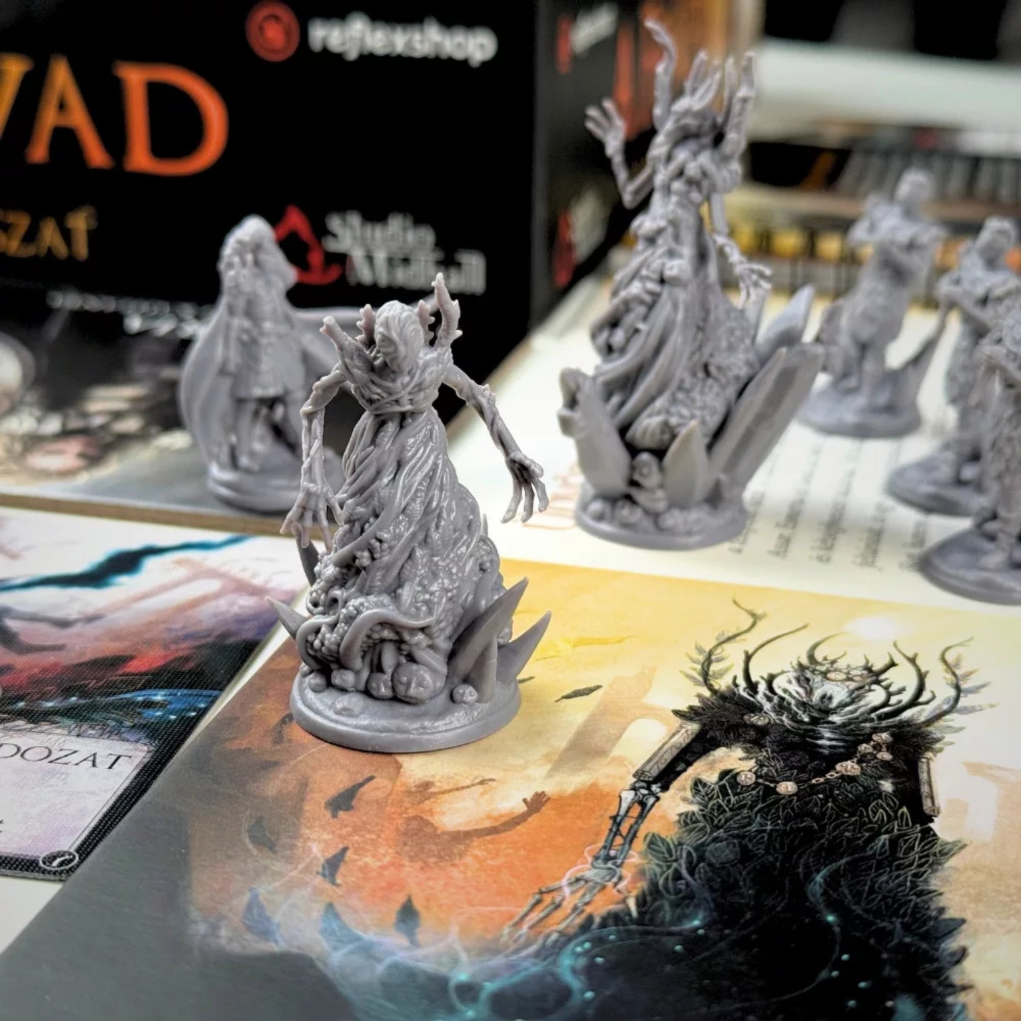 A Fenevad: A Nagy Vadászat társasjáték részletes szürke miniatűr figurái közelről, karakterlapok és illusztrált játékelemek előtt.