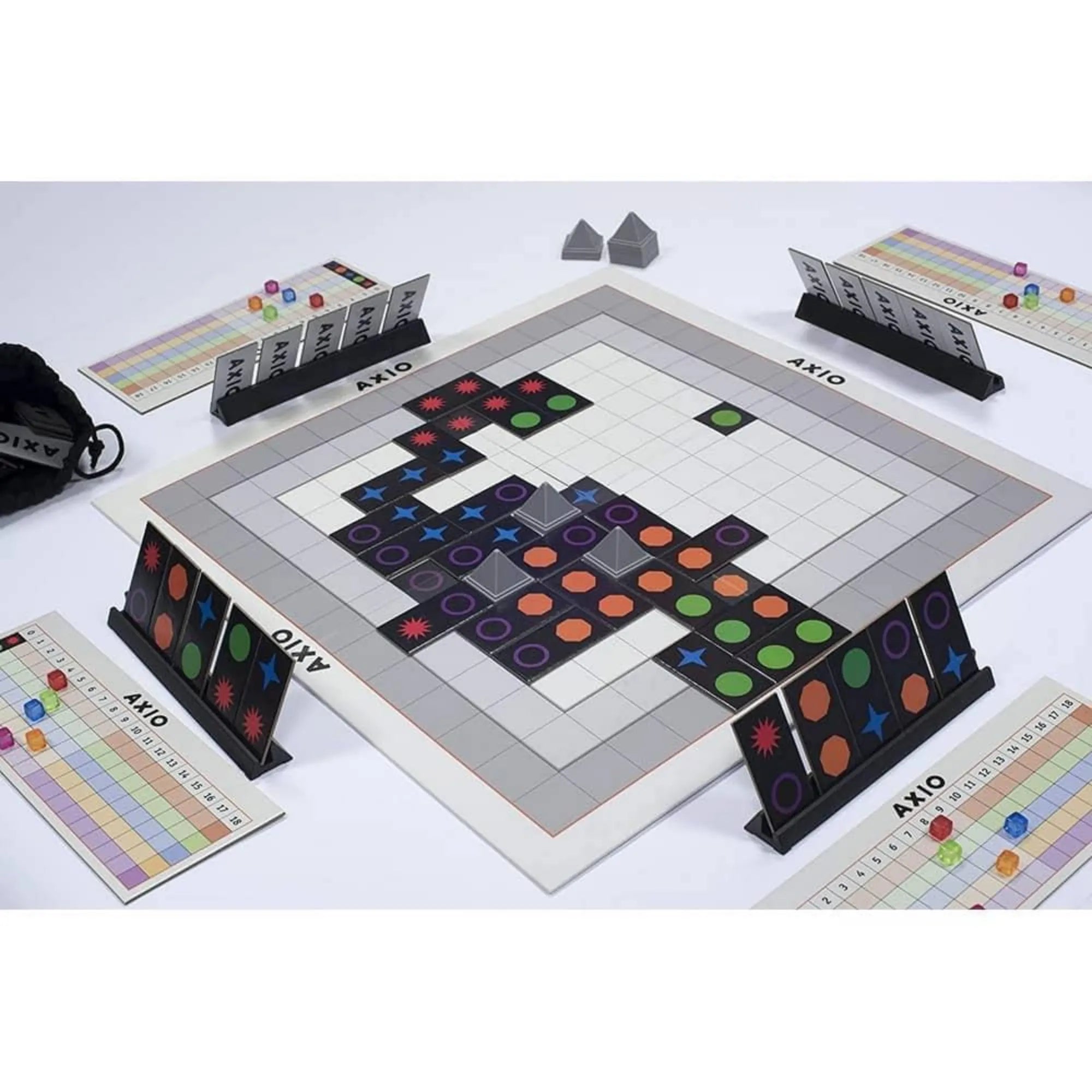 AXIO logikai társasjáték játék közben, központi játéktáblával, színes szimbólumos lapkákkal, pontozótáblákkal és játékos paravánokkal, felülnézeti elrendezésben