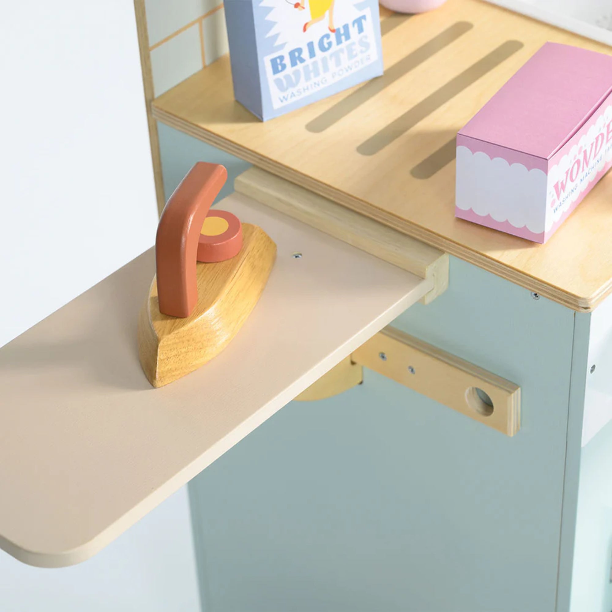 Fa vasaló egy kihúzható vasalódeszkán, mellette színes mosóporos dobozok és kiegészítők, pasztell árnyalatú mosoda felületen.