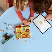 Gyerek és szülő közösen építi a LEGO Bluey 11203 házat egy világoskék asztalon. Az építés során a nyitott házrész jól látható, különböző elemek, minifigurák, kiegészítők és egy tablet is jelen van, amin az utasításokat követik digitálisan.