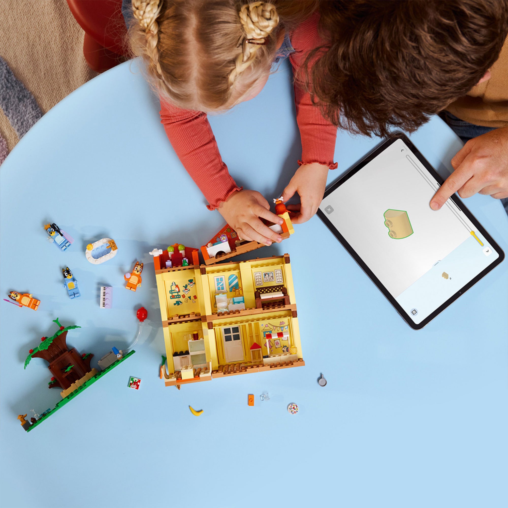 Gyerek és szülő közösen építi a LEGO Bluey 11203 házat egy világoskék asztalon. Az építés során a nyitott házrész jól látható, különböző elemek, minifigurák, kiegészítők és egy tablet is jelen van, amin az utasításokat követik digitálisan.