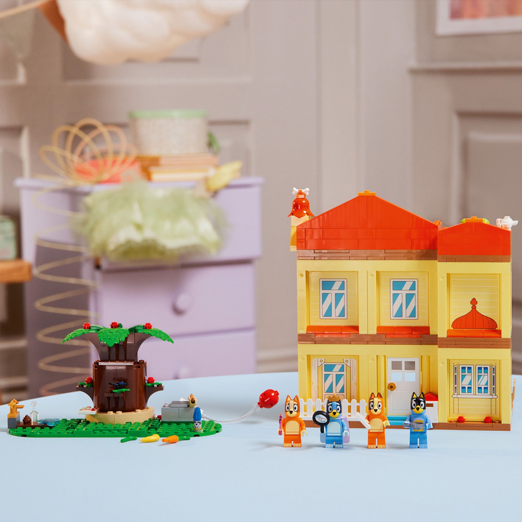 A LEGO Bluey családi ház hátsó, zárt homlokzata, élénk sárga falakkal, kék ablakokkal és piros tetővel. Az előtérben Bluey, Bingo, Bandit és Chilli minifigurák állnak, mögöttük a nagy lombos fa, lufi és egy kerti jelenet. A szoba háttérben világos, otthonos környezetben készült.