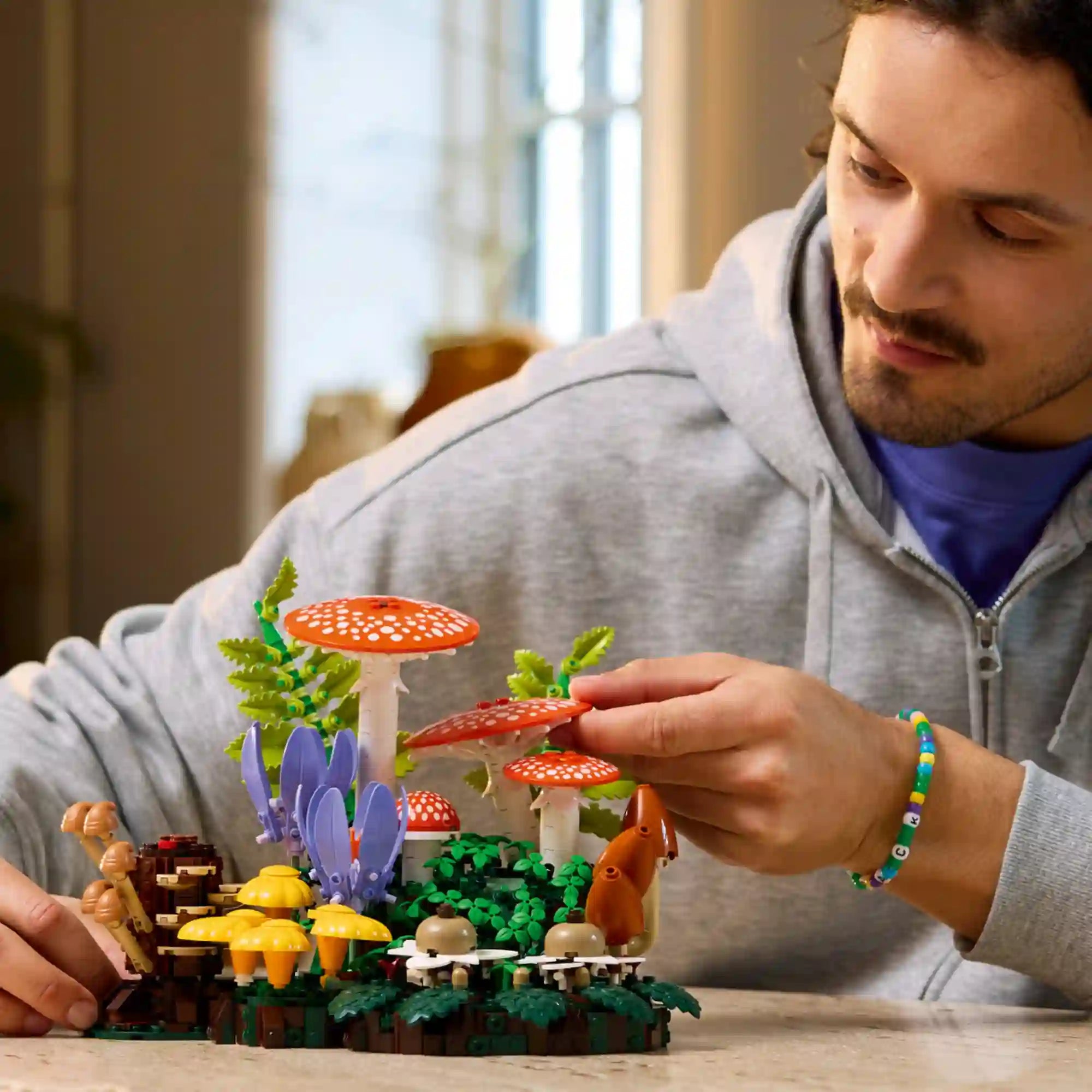 LEGO Botanicals Erdei gombák (11505) építőkészlet életkép, amelyen egy építő a részletgazdag erdei jelenetet állítja össze, légyölő galócákkal, páfrányokkal és színes virágokkal.