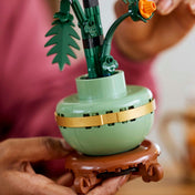 Közeli kép a LEGO Botanicals 10368 Chrysanthemum zöld kaspójáról, arany színű díszítéssel, miközben egy kéz tartja az építményt.