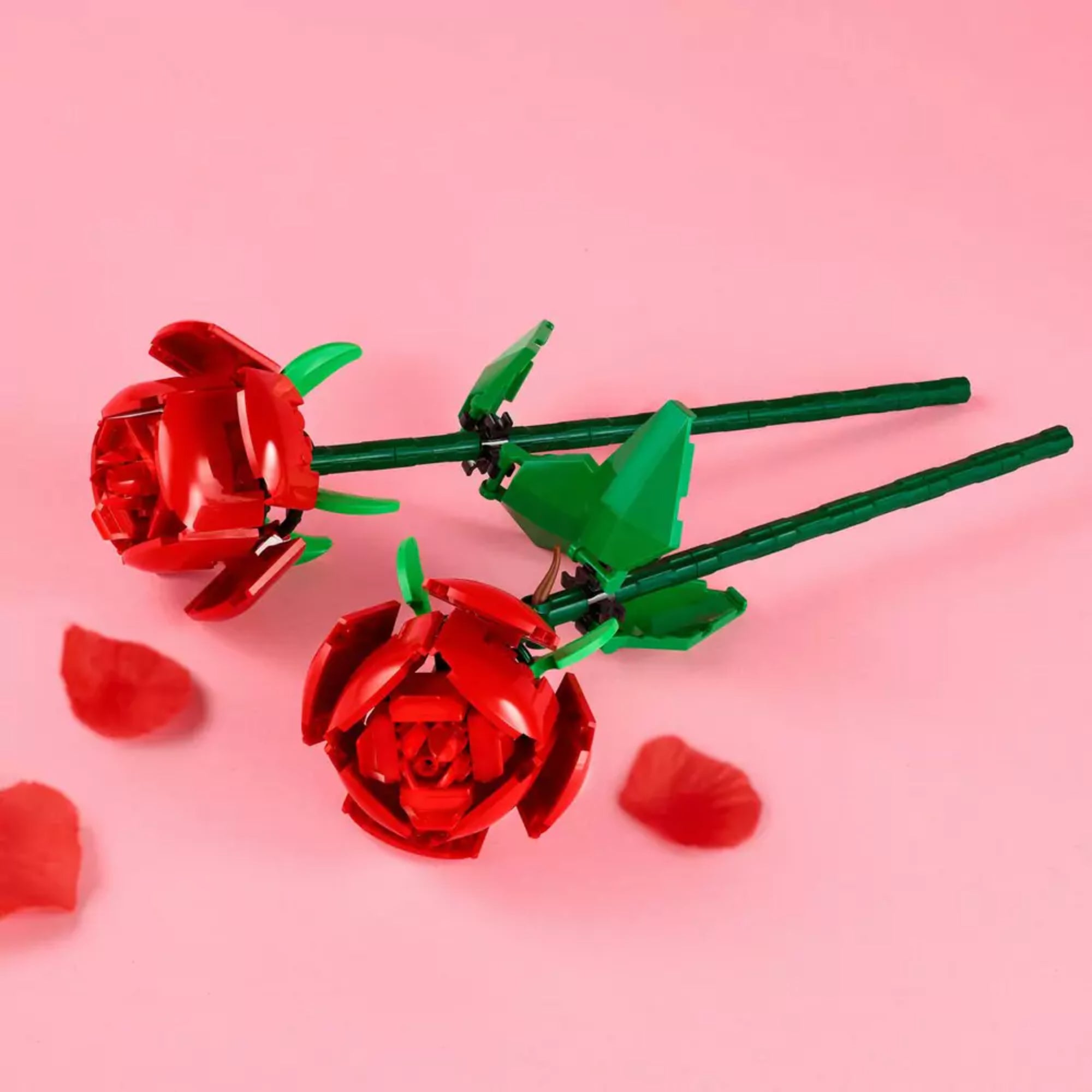 Két piros LEGO rózsa zöld szárral és levelekkel elhelyezve rózsaszín háttéren, a LEGO Rózsák (40460) készlet elemeiként, szétszórt sziromdíszítéssel.