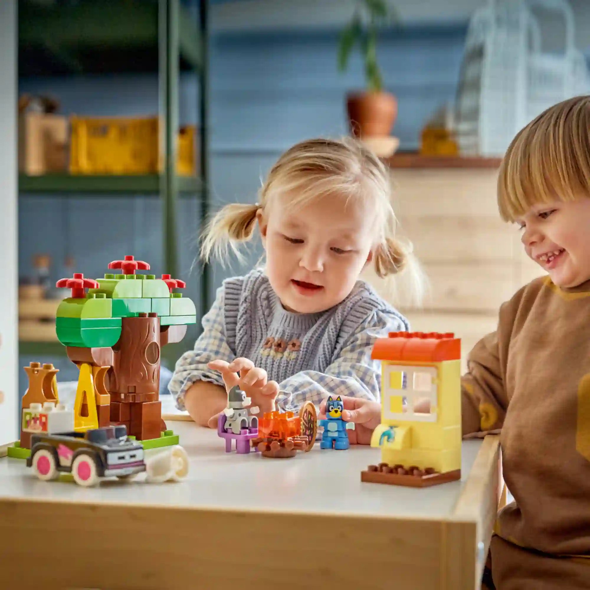LEGO DUPLO Bluey: Pizza parti Muffinnal és Bluey-val (10469) használat közben, két kisgyermek által játszott jelenettel, fával, házrésszel, autóval, pizzával és Bluey karakterfigurákkal.