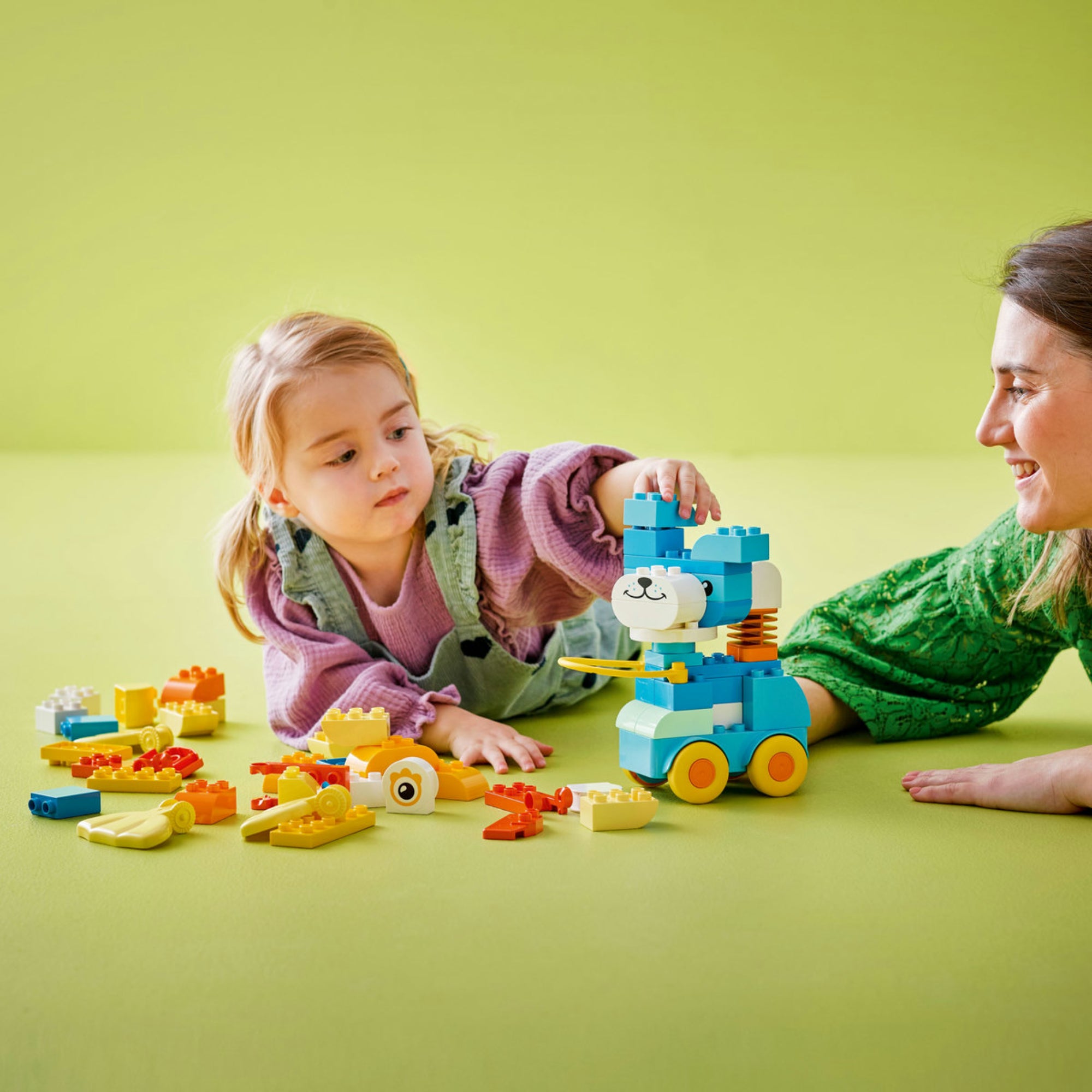Egy kislány és egy nő együtt játszanak a LEGO DUPLO 10448-as készlettel egy zöld háttér előtt. A kislány egy világoskék és fehér medvefigurát helyez rá a guruló alapra, körülöttük sok színes építőkocka található.