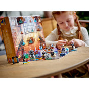 Kislány játszik a LEGO Harry Potter adventi naptár elemeivel egy asztalon, háttérben a kibontott naptár és az elrendezett figurák, köztük Harry, Voldemort és különféle varázslatos jelenetek.