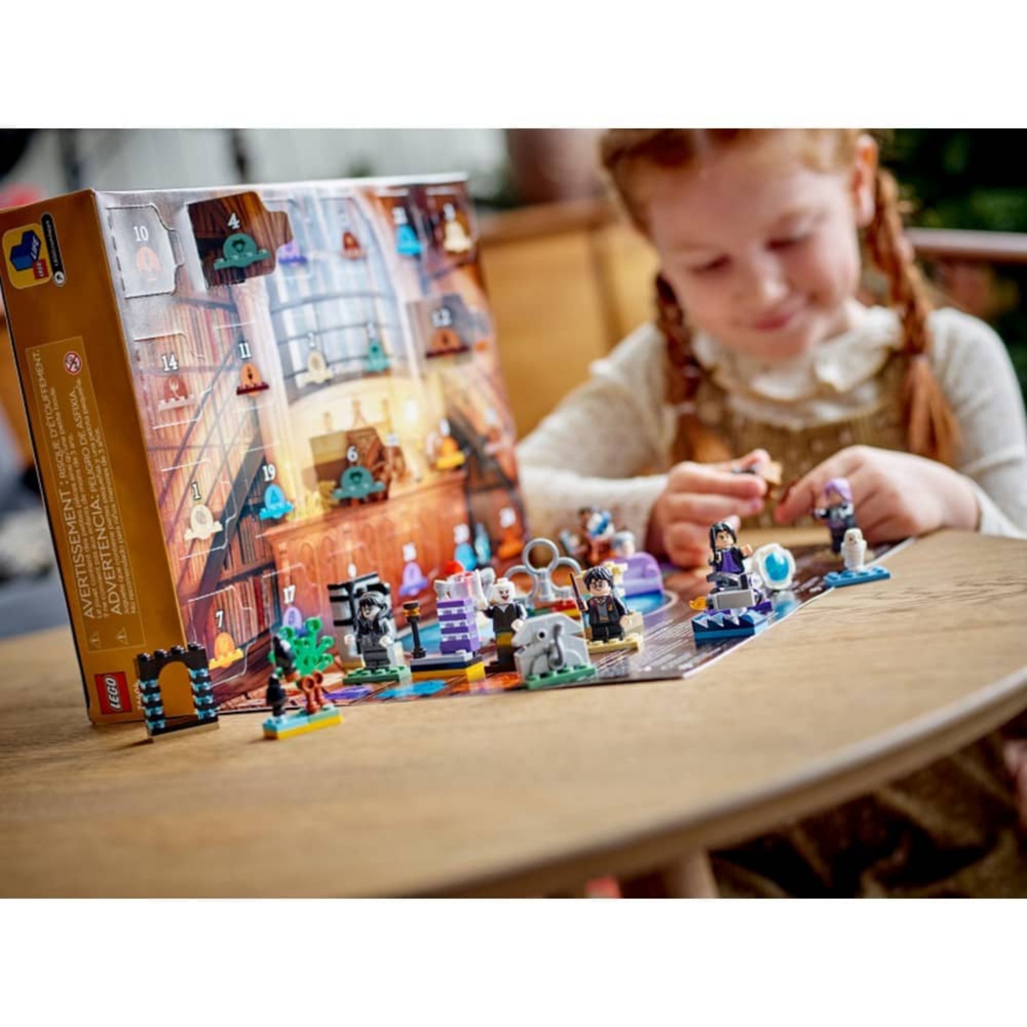 Kislány játszik a LEGO Harry Potter adventi naptár elemeivel egy asztalon, háttérben a kibontott naptár és az elrendezett figurák, köztük Harry, Voldemort és különféle varázslatos jelenetek.
