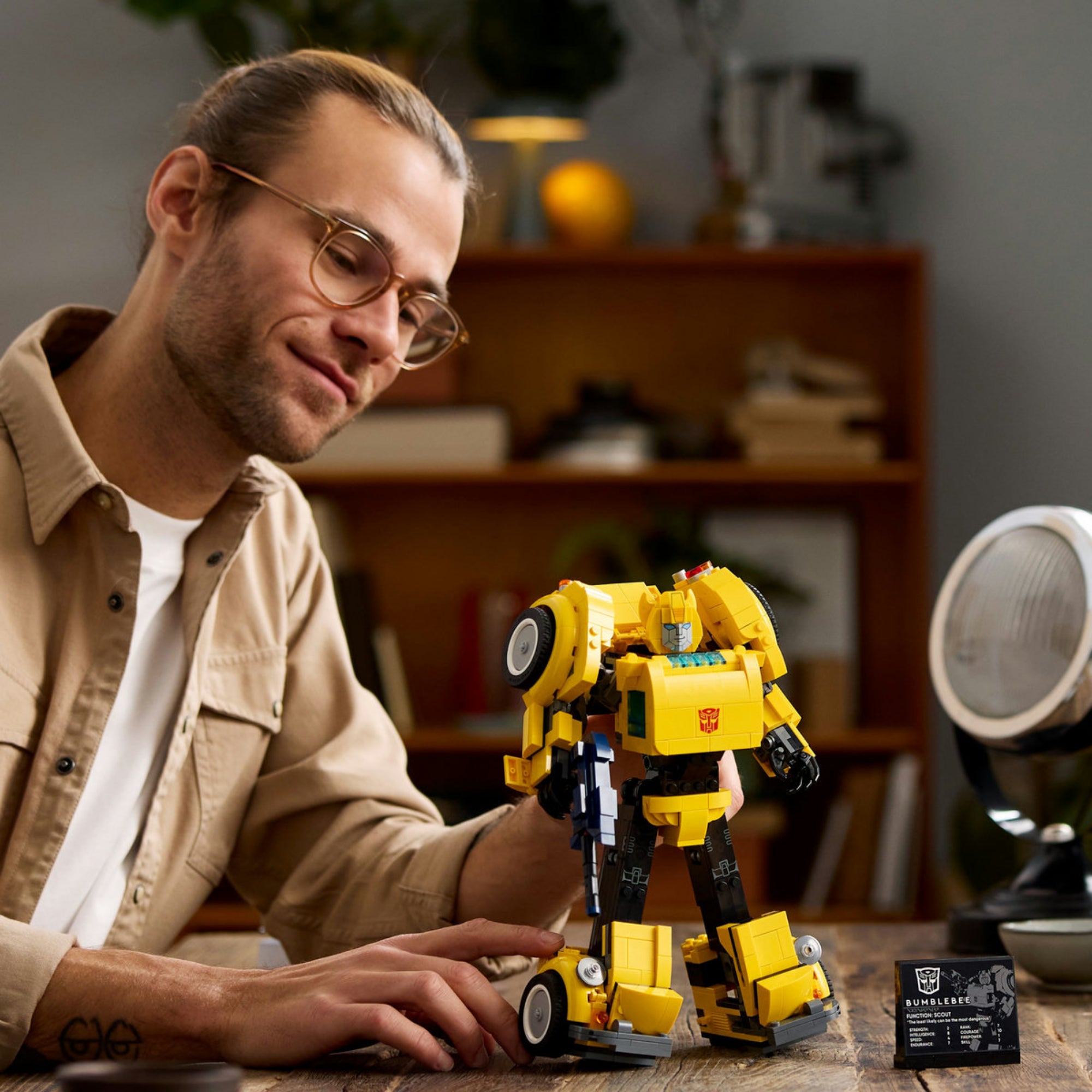 Egy felnőtt építő a LEGO Icons Bumblebee robotot mutatja be asztalon, mellette az információs tábla, háttérben otthoni környezet.