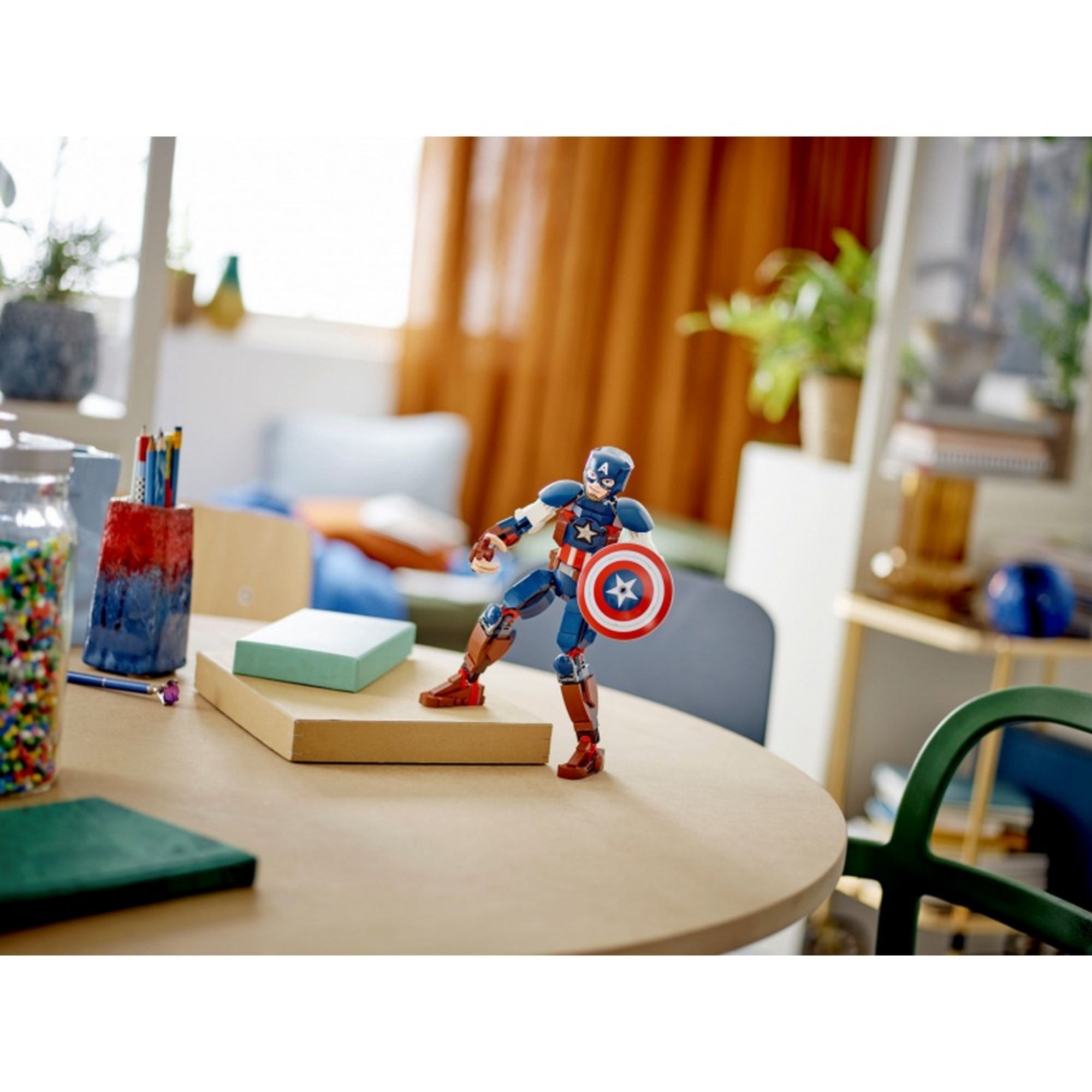 Az Amerika Kapitány LEGO figura kiállítva egy gyerekasztalon, háttérben otthoni környezettel, a figura akciópozícióban, pajzzsal a kezében.
