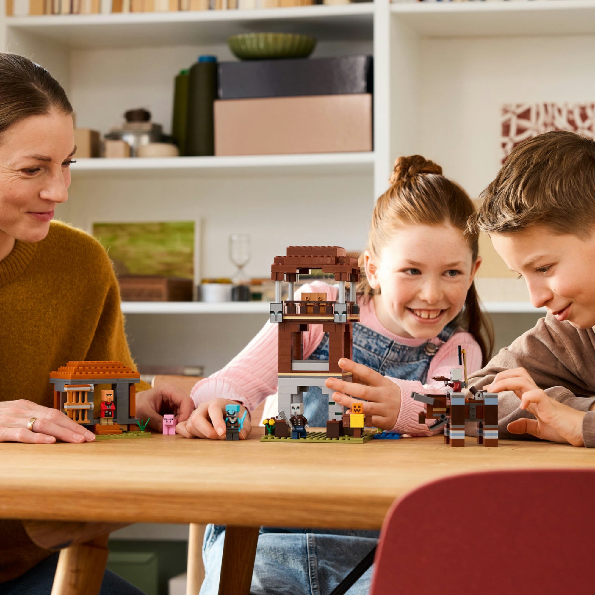 Gyerekek és egy felnőtt közösen játszanak a LEGO Minecraft A Fosztogató őrtorony és a Romboló (21278) készlettel egy asztalon, mosolygó arccal, miközben figurákat mozgatnak.