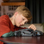 Egy fiú játszik a LEGO Technic Koenigsegg Jesko modellel, miközben mozgatja a jármű elemeit; a háttérben egy otthonos nappali látható.