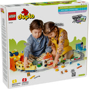 Két kisgyerek játszik a LEGO DUPLO 10428 vonatkészlettel, miközben sínpályán vezetik a sárga mozdonyt; a háttérben láthatók az állomás, akciókockák és egyéb színes építőelemek is.