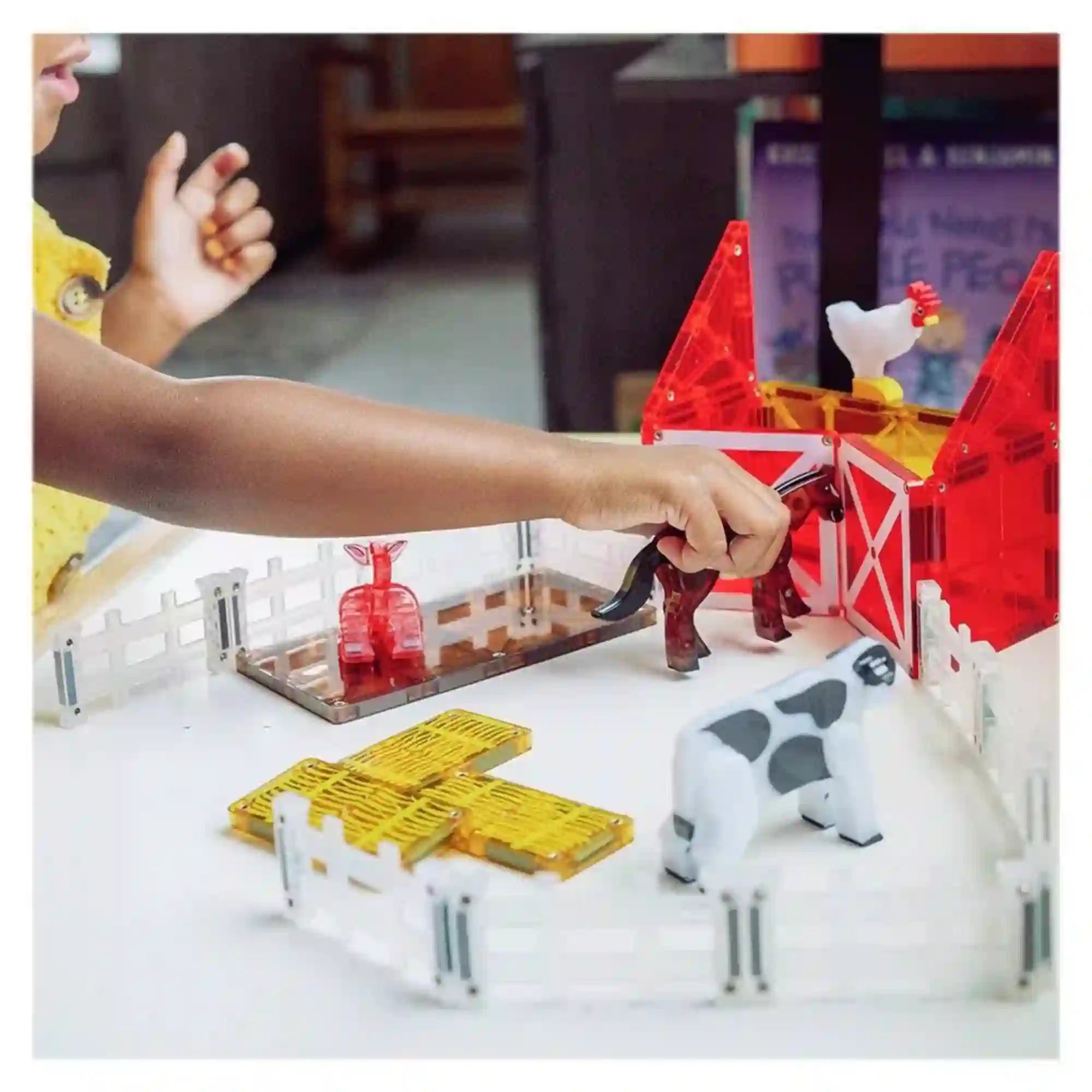 MAGNA-TILES Farm Animals mágneses építőkészlettel játszó gyermek, aki egy barna lovat mozgat a piros istálló és a fehér kerítések között.