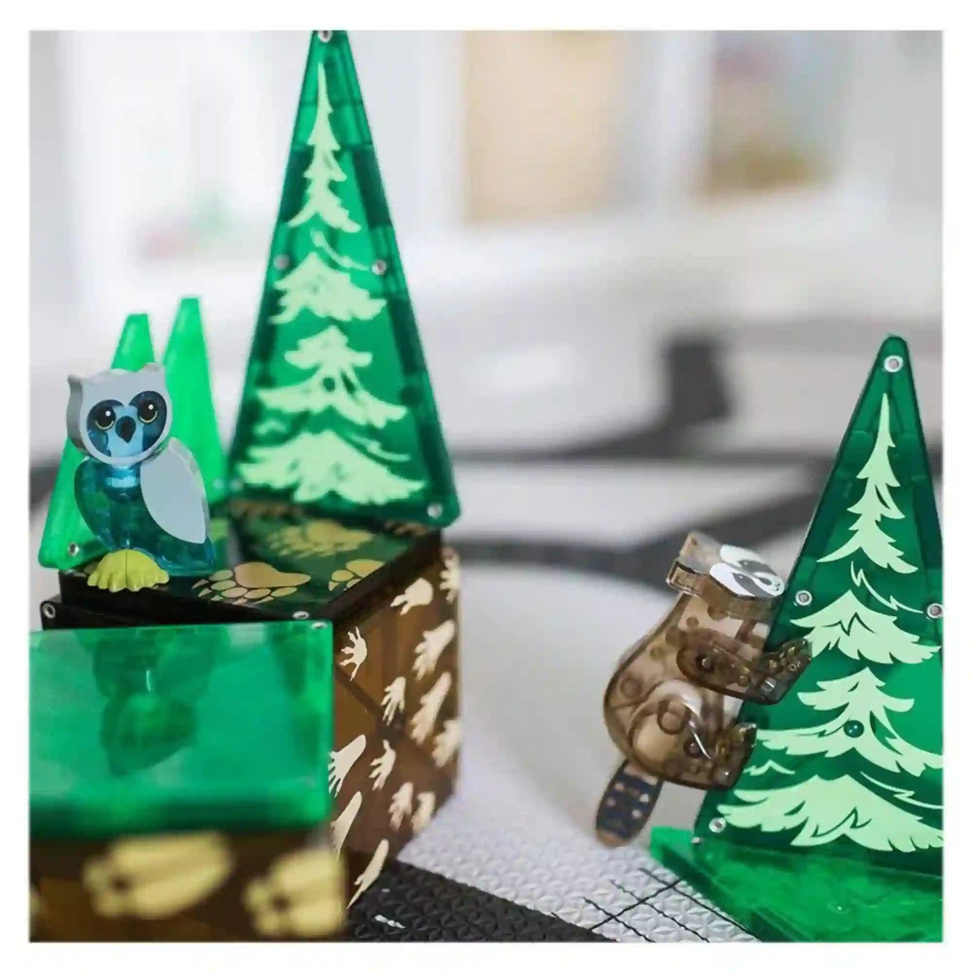 MAGNA-TILES Forest Animals mágneses építőkészlet erdei jelenete, kék bagollyal, fára kapaszkodó mosómedvével és zöld fenyőfákkal.