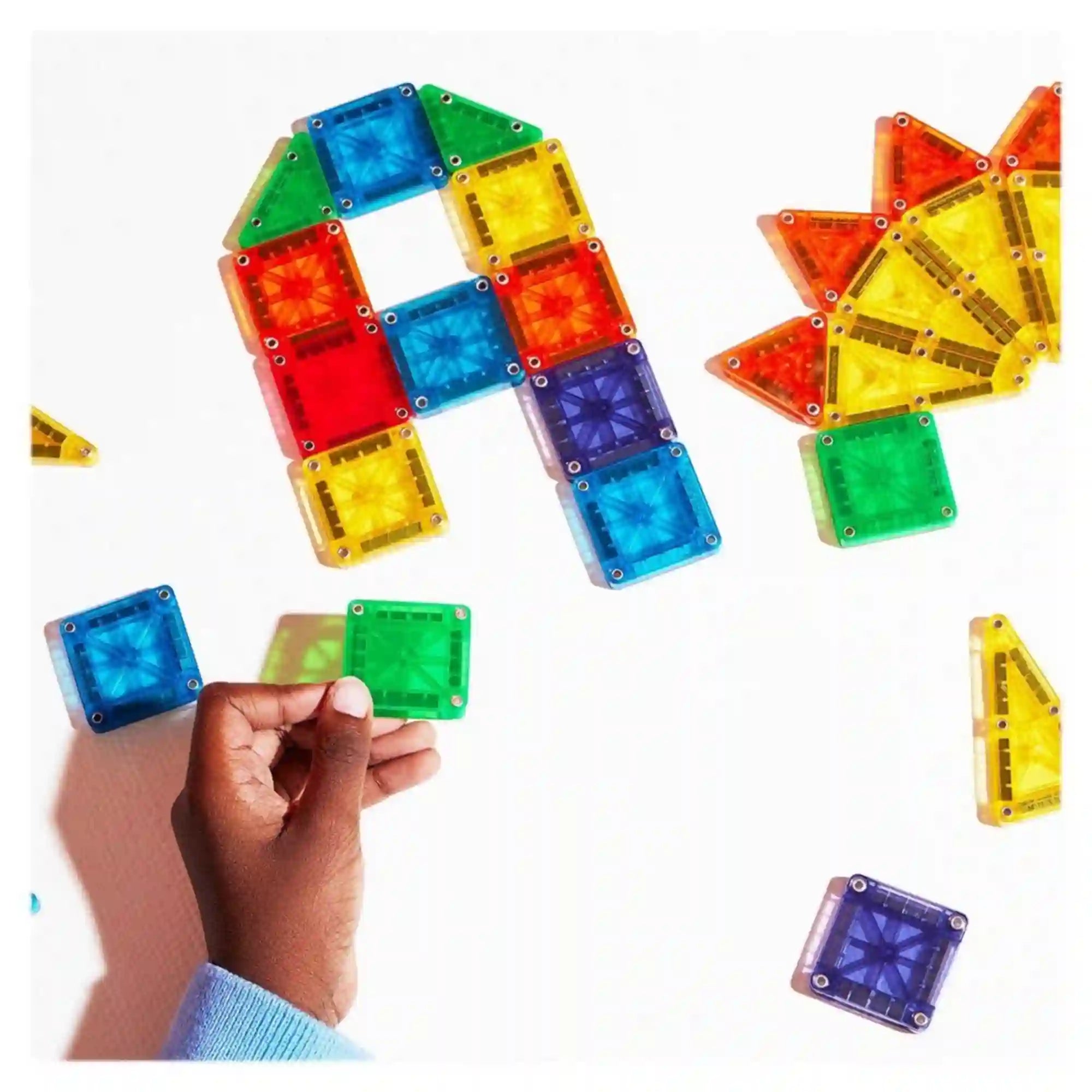 MAGNA-TILES microMAGS 70 darabos építőkészlet áttetsző, színes mágneses lapjai fehér felületen, egy gyermek kezében zöld négyzet alakú építőelemmel.