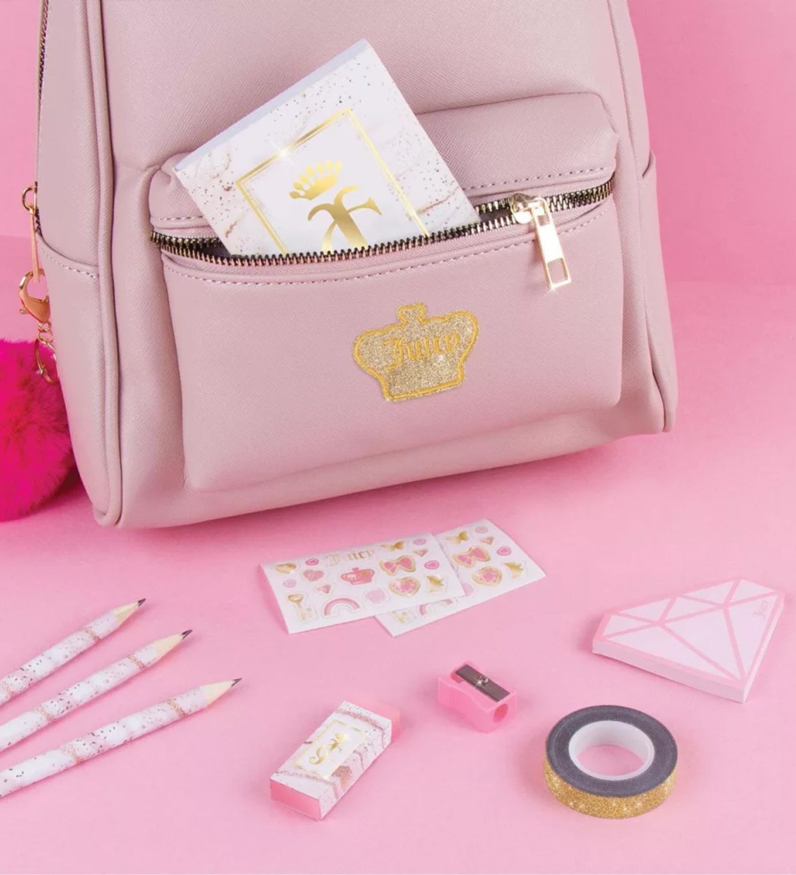 Juicy Couture Deluxe Stationery Set elemei rózsaszín háttéren, füzet a táskában, ceruzák, radír, hegyező, matricalap és arany dekortapasz elrendezve.
