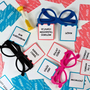 Nagyi bugyiban társasjáték játékelemei közelről: színes műanyag szemüvegekbe helyezett szerep- és szitukártyák, feliratos kártyák és homokóra játék közbeni elrendezésben.