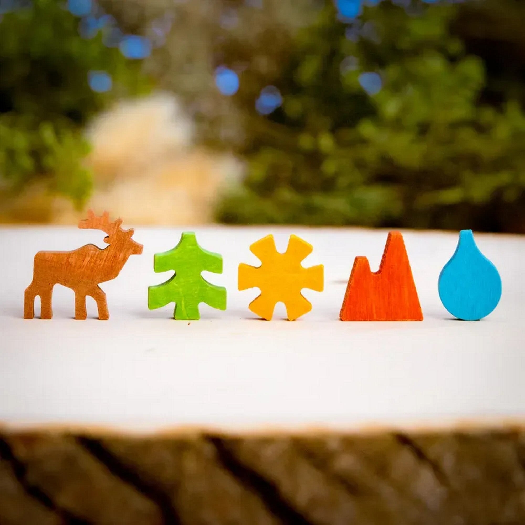 Parks társasjáték fa játékelemek, színes figurák sorban (szarvas, fa, nap, hegy, vízcsepp) természetes háttér előtt.