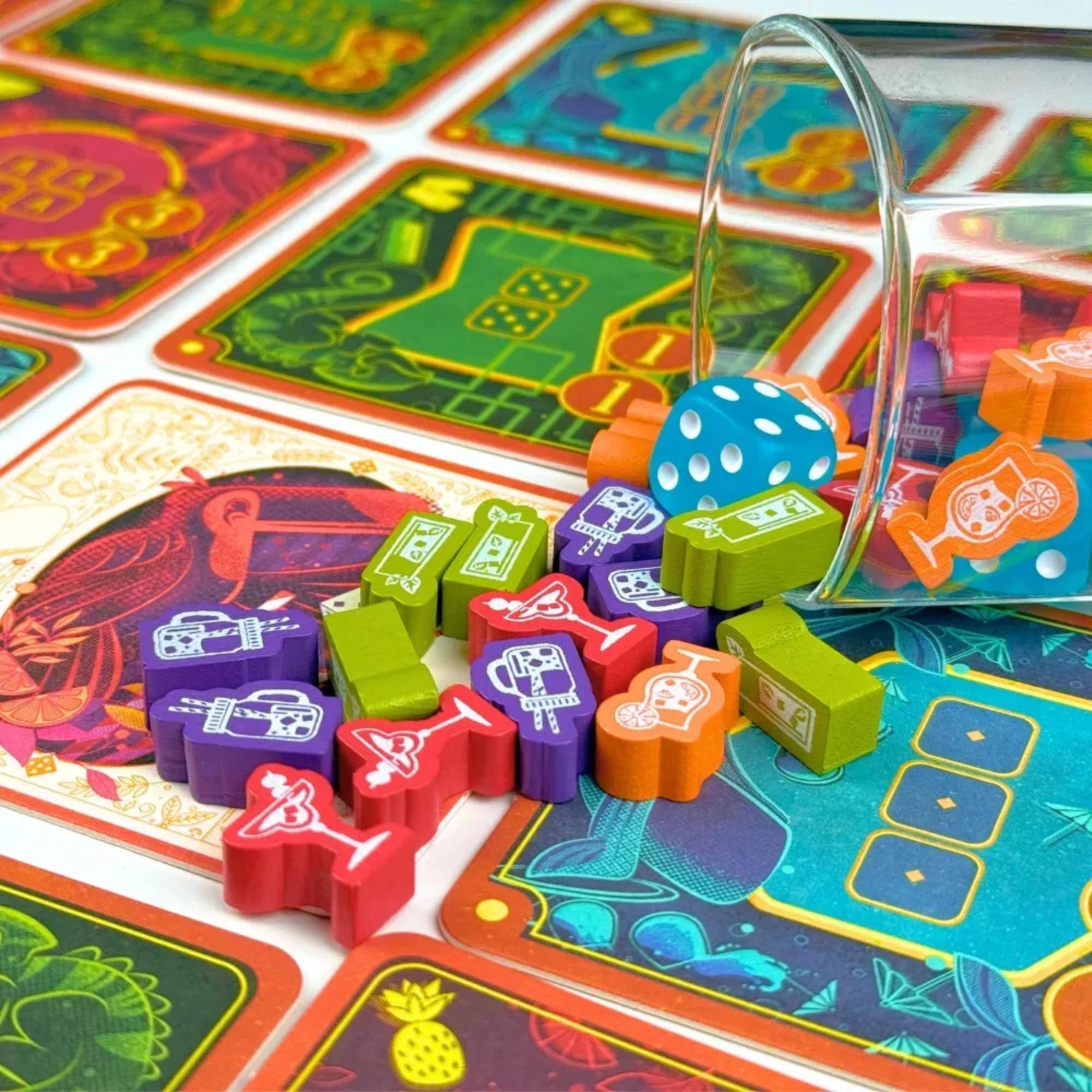 Közeli részlet a Piña Coladice játékelemekről: kiömlött színes fa koktélikonok, türkiz dobókocka és élénk mintázatú játékalátétek a háttérben.