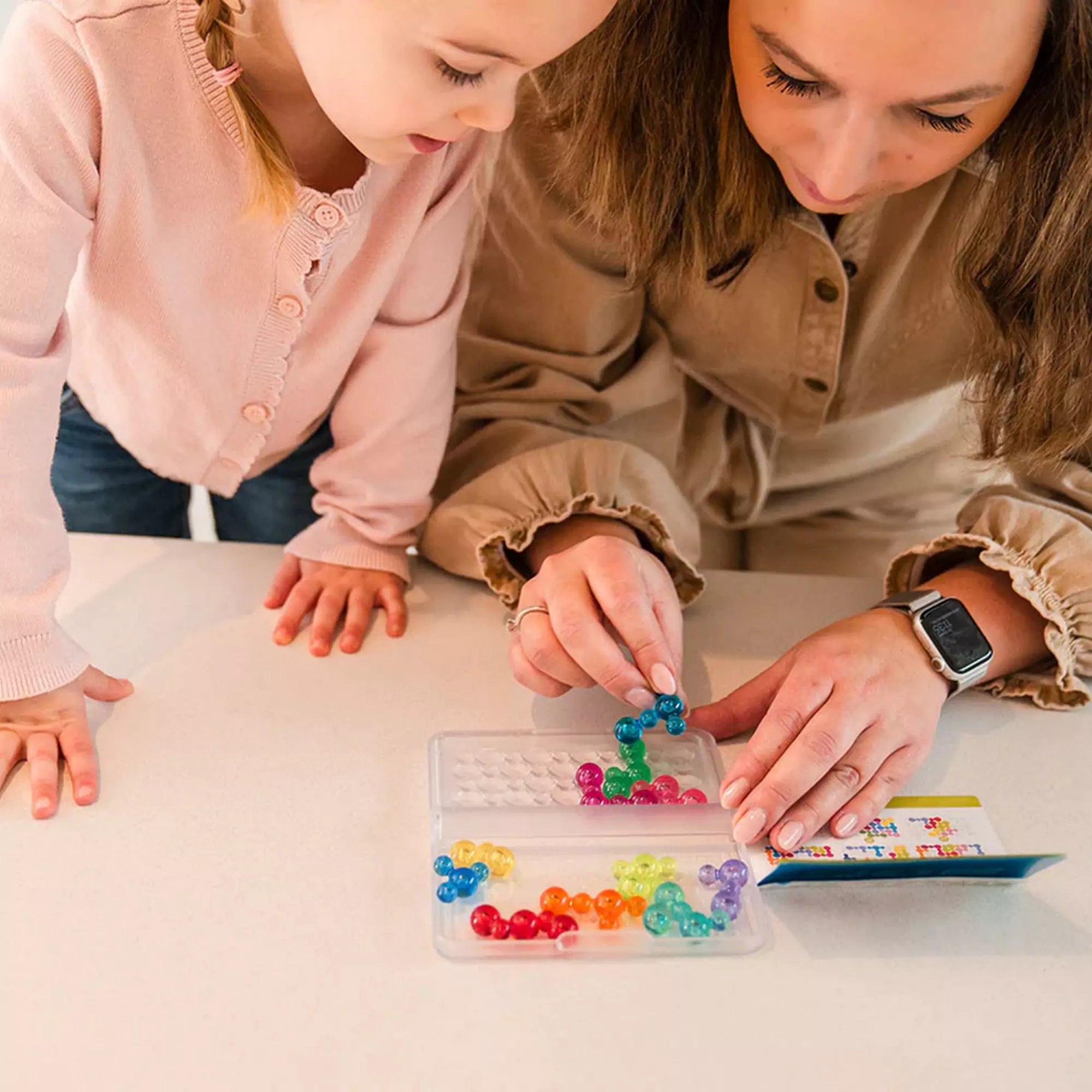 Smart Games IQ Bubbles. Egy felnőtt és egy gyermek együtt játszik az IQ Bubbles játékkal, a felnőtt segít elhelyezni az egyik kék buborékformát.