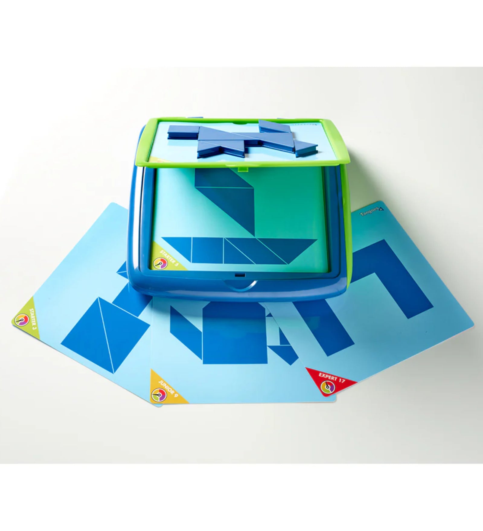 A képen a Smart Games Tangoes Junior logikai játék használat közben látható. A középen elhelyezkedő mágneses játéktáblán geometriai formákból álló kirakós feladat látható, amelyet a gyerekeknek kell megoldaniuk a hozzá tartozó kék tangram elemek segítségével. A tábla alatt több kihíváskártya található, különböző nehézségi szintekkel, például „Junior 9” és „Expert 17”. A színes sarokjelölések segítenek megkülönböztetni a kártyák nehézségi szintjét. 
