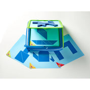 A képen a Smart Games Tangoes Junior logikai játék használat közben látható. A középen elhelyezkedő mágneses játéktáblán geometriai formákból álló kirakós feladat látható, amelyet a gyerekeknek kell megoldaniuk a hozzá tartozó kék tangram elemek segítségével. A tábla alatt több kihíváskártya található, különböző nehézségi szintekkel, például „Junior 9” és „Expert 17”. A színes sarokjelölések segítenek megkülönböztetni a kártyák nehézségi szintjét. 