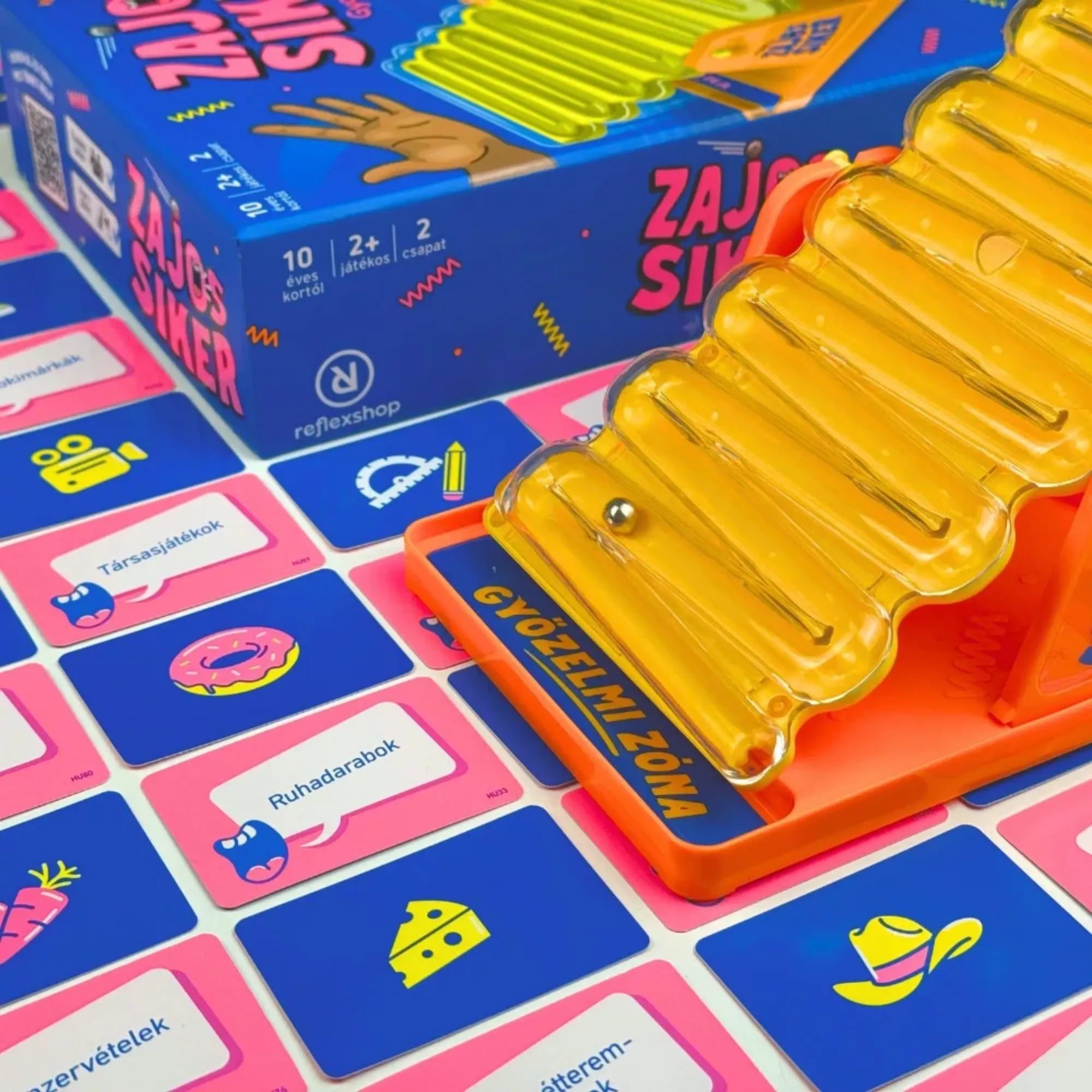 Zajos siker társasjáték játék közben, sárga zajkeltő pályával és guruló golyóval, körülötte elhelyezett feladatkártyákkal, pörgős ügyességi és kommunikációs partijáték bemutatása.