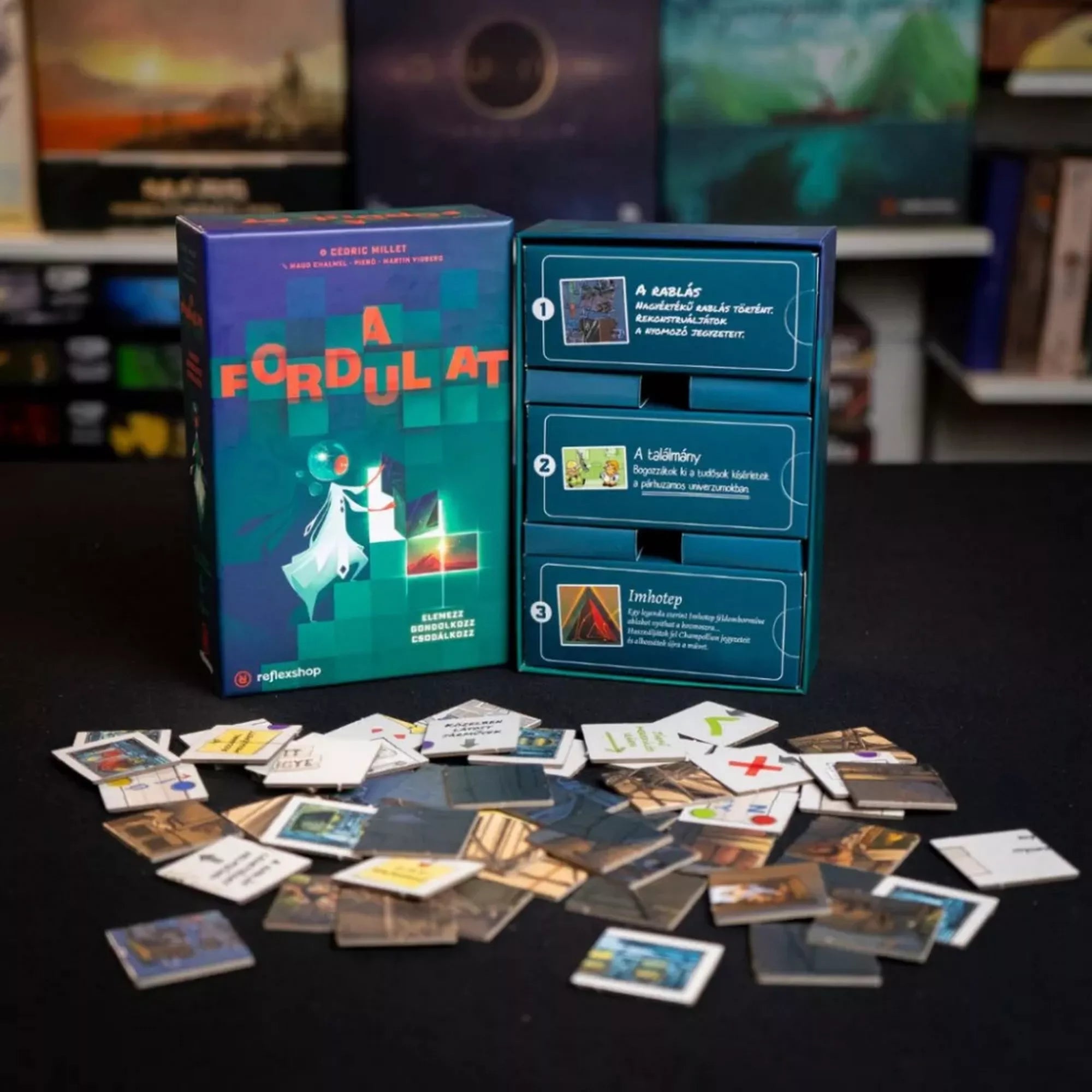 A Fordulat társasjáték doboza és a három külön játékrész a kiborított lapkákkal együtt egy sötét játéktéren. A doboz mellett a rejtvénydobozok jól láthatók.