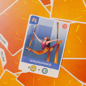 Az Évad társasjáték levegőakrobata kártyája közelről, narancssárga hátlapos kártyákon, a művész szövetfüggönyökön függeszkedve.