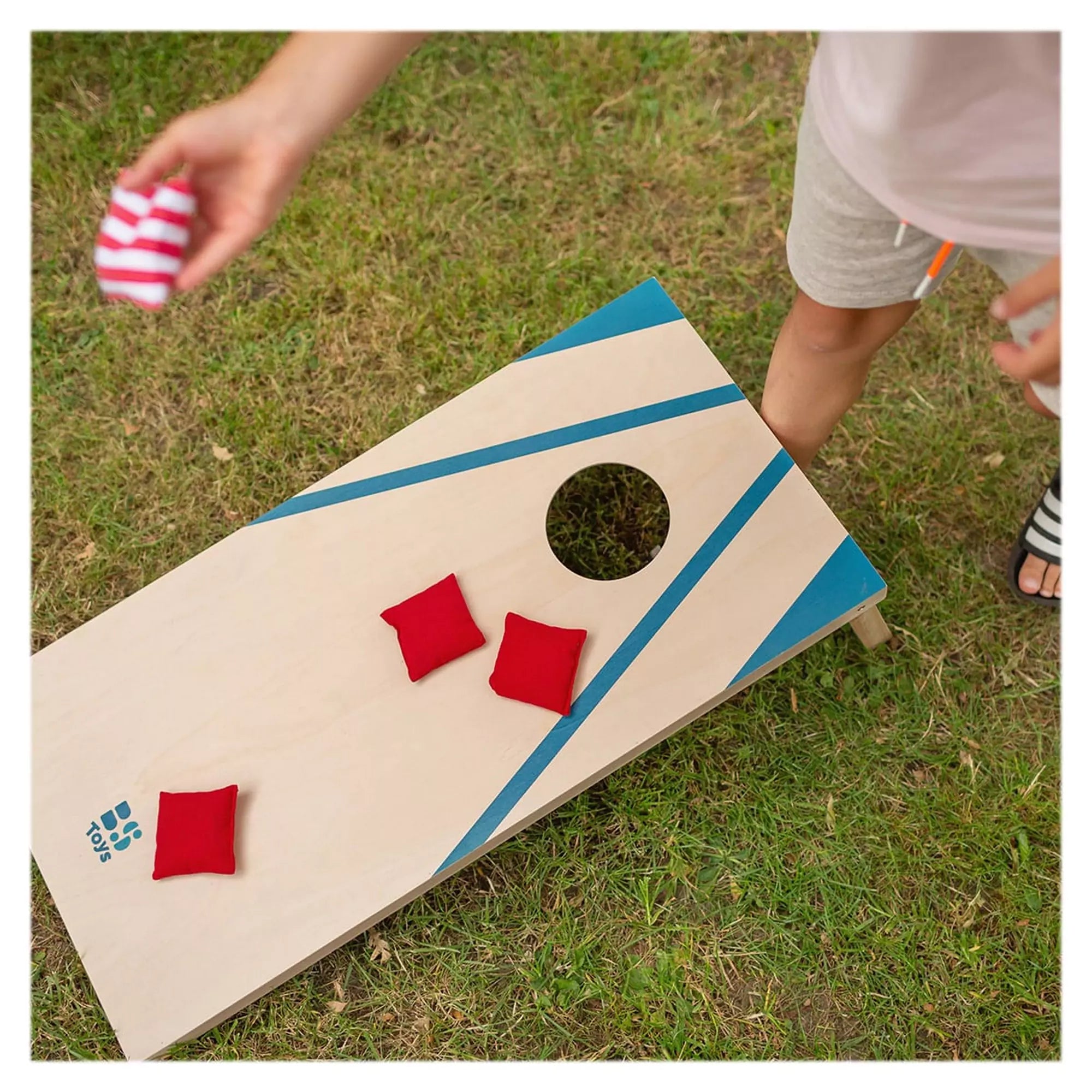 Felülnézetből látható, ahogy egy gyermek piros és csíkos zsákokat dob a BS Toys Cornhole Set kék mintás táblájára.