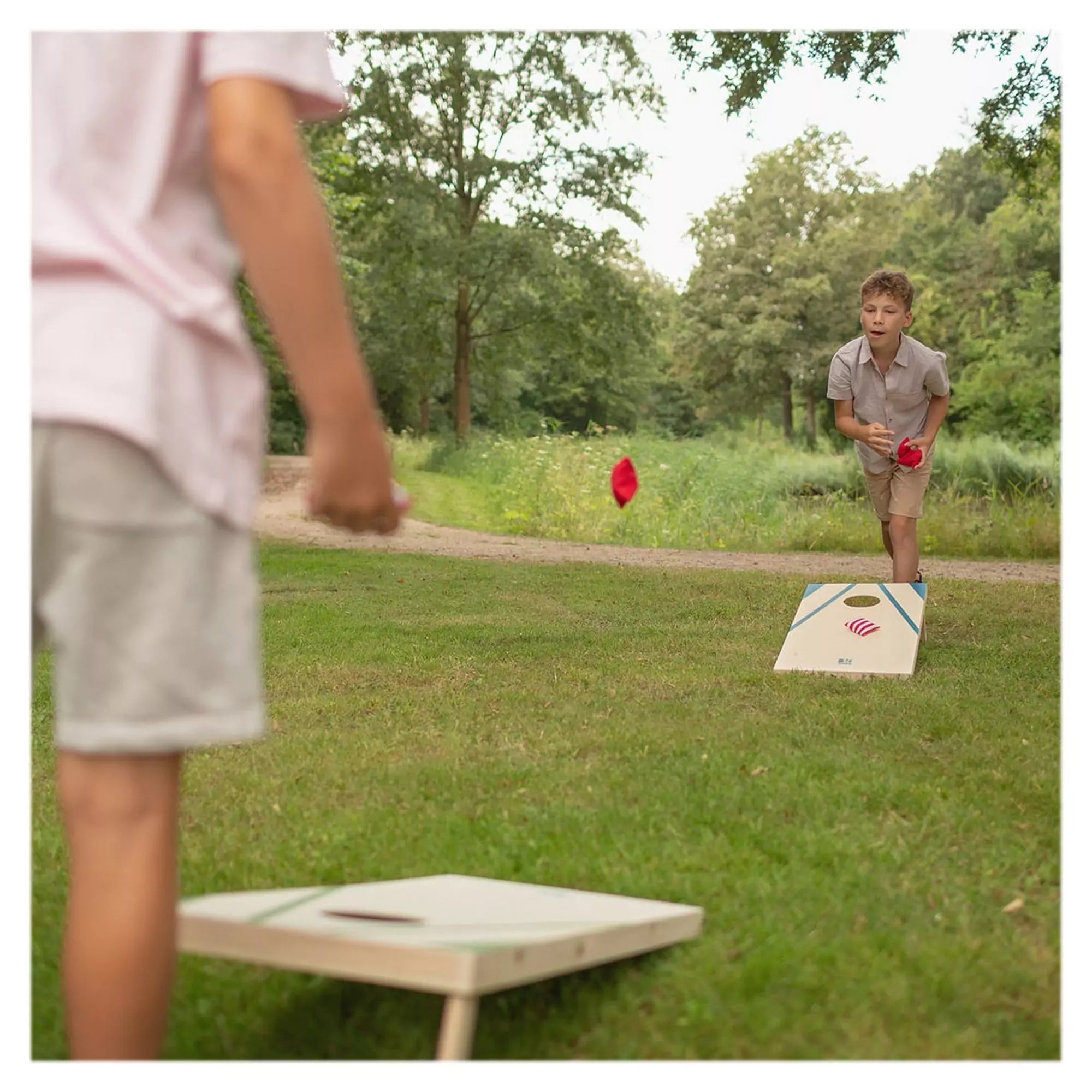 Két gyerek játszik a füvön a BS Toys Cornhole Set játékkal, egyikük éppen zsákot dob a tábla felé.