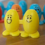 Két sárga Balance Beans figura összekapcsolva, mosolygós arccal, fapadlón állnak, háttérben további színes figurák homályosan.