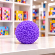 Cool Mix Squishy bolyhos tapintású labda 6 cm stresszlevezető játék (lila), lila színű, rücskös felületű labda játékbolt pultján, elmosódott polcok előtt.