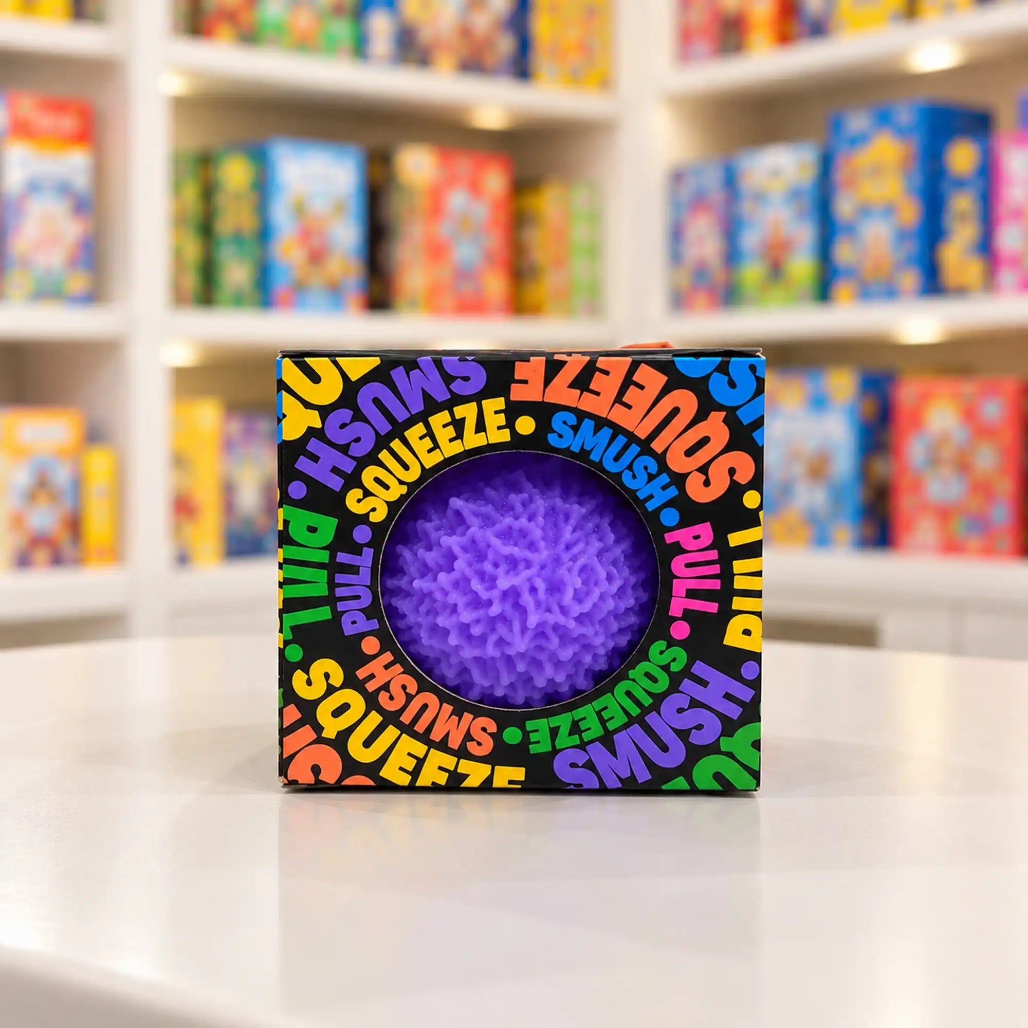 Cool Mix Squishy: Bolyhos tapintású labda 6 cm stresszlevezető játék (lila), fekete, színes feliratokkal díszített dobozban, kerek ablakon keresztül látható lila labdával.